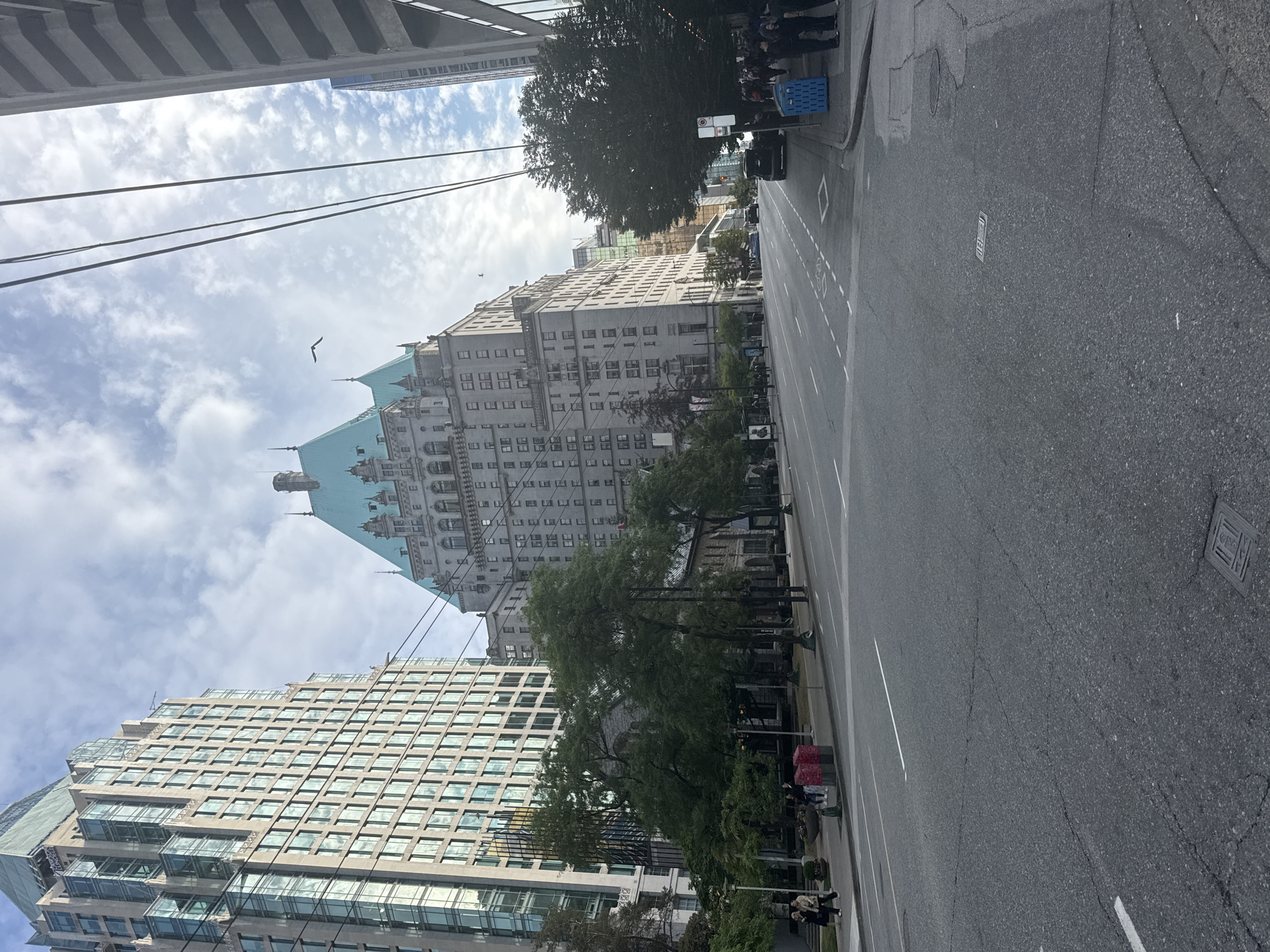 This is a daytime, eye-level shot of a city street in Vancouver, Canada, featuring several prominent buildings and urban infrastructure. The image is dominated by the Fairmount Hotel Vancouver, a large, historic building with distinctive turquoise roofs and multiple spires, situated in the center of the frame. To the left of the Fairmont Hotel, a modern, tall skyscraper with a glass facade rises, its windows reflecting the sky. Another tall, gray building with a repeating pattern of windows is visible on the far right.
The street in the foreground is paved with asphalt and marked with white lane dividers. A few cars are visible on the street, including a dark SUV on the right side. Sidewalks line both sides of the street, with pedestrians walking along them. On the left sidewalk, a few people are walking and standing near some trees. On the right sidewalk, a group of people are gathered near some street furniture, including a blue recycling bin.
Trees with green foliage are scattered along the sidewalks, adding a touch of nature to the urban landscape. The sky is partly cloudy with some patches of blue sky visible. Overhead, power lines or communication cables crisscross the sky. The overall impression is that of a bustling urban environment with a mix of historical and modern architecture.