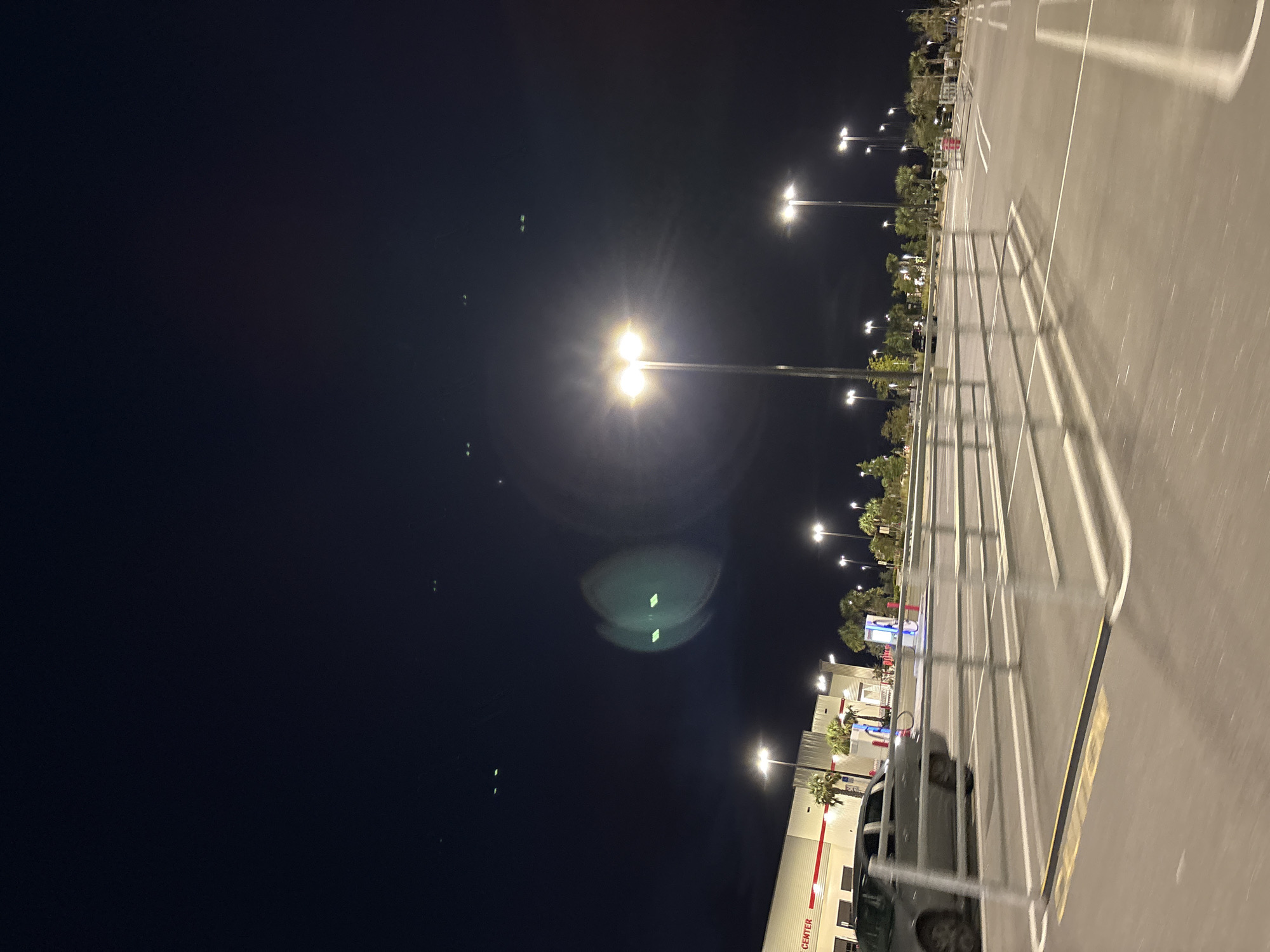 The image depicts a nighttime scene at a Costco store in North Port, Florida. The foreground shows an empty asphalt parking lot with white lane markings and yellow directional arrows. A silver sedan is parked on the left side of the frame. A metal railing system, likely for queueing, is also visible in the parking lot.
In the background, the Costco building is illuminated, with its signage visible. Palm trees are planted in front of the building. Several streetlights with bright lamps illuminate the parking lot. The sky is dark, with a few distant lights that could be stars or distant aircraft. There is a noticeable lens flare or light artifact in the sky to the right of the center, appearing as two bright green squares with a blurry circular halo. Several other smaller green lights are scattered across the sky.