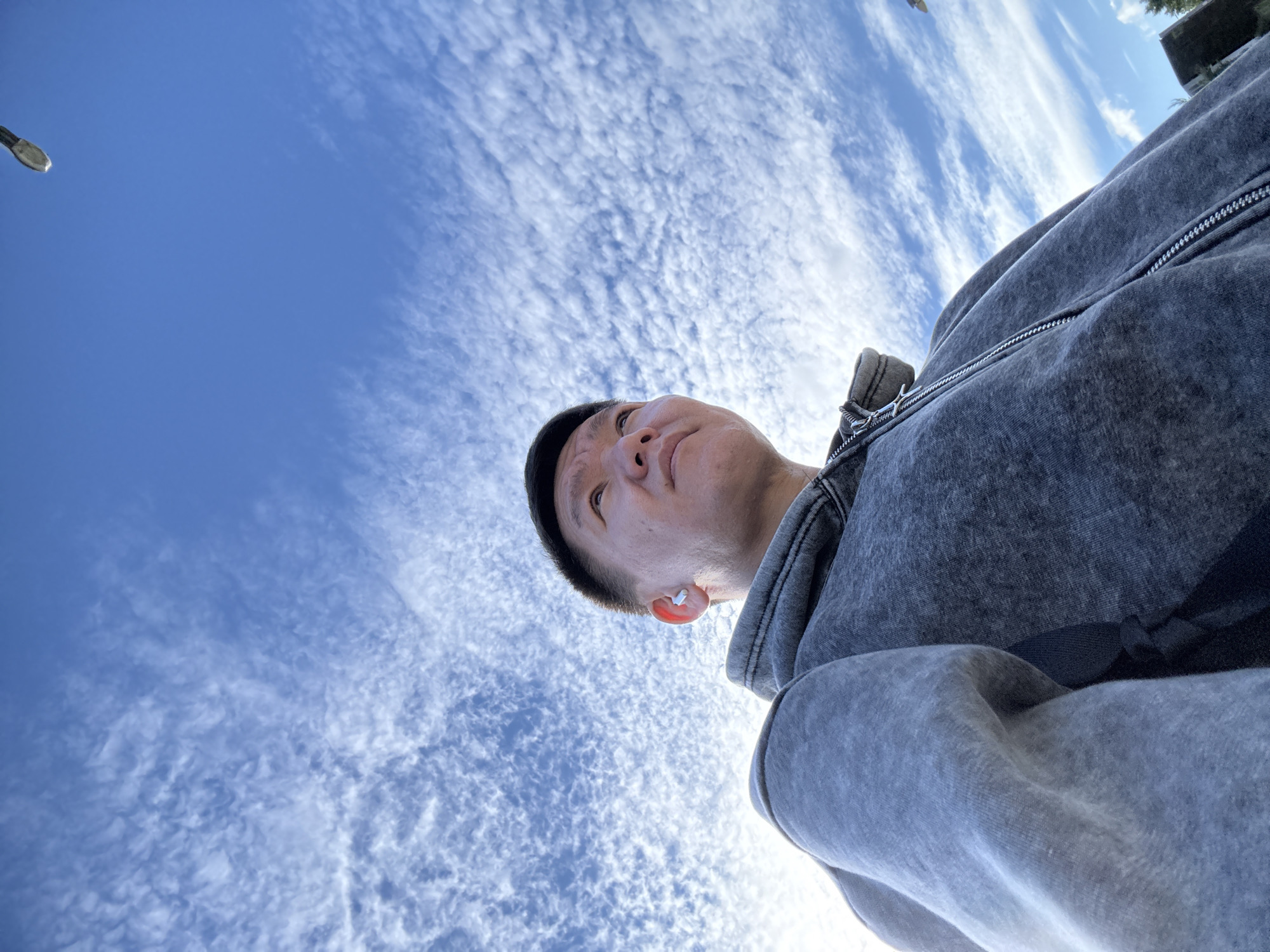 A low-angle, medium shot captures a person from the chest up, looking up at the sky. The person, appearing to be of Asian descent, has short dark hair and is wearing a grey zip-up hoodie and a small white earbud. The sky above is a vibrant blue, filled with wispy, elongated white clouds that stretch across the frame, creating a textured, almost painterly effect. In the upper right corner of the image, a distant street lamp is visible against the bright sky. The background also shows a hint of greenery on the right side, suggesting an outdoor setting, possibly a park or open area within a city. The lighting suggests it is daytime, with the sun likely positioned behind the viewer or to the left, casting a gentle illumination on the person's face and the clouds. The overall mood is calm and contemplative, with the upward gaze of the subject drawing attention to the expansive sky.