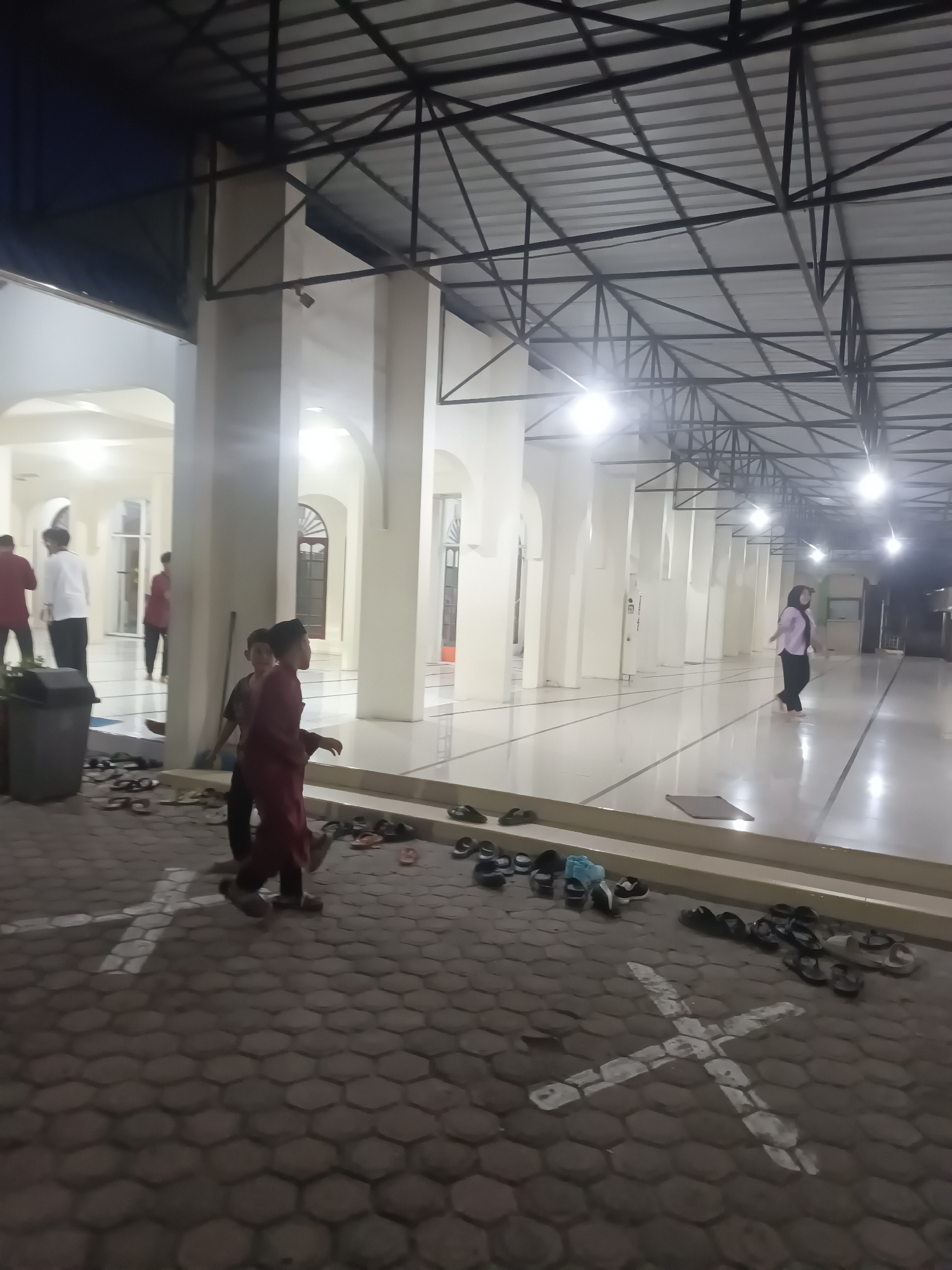 The image shows a view of a mosque's outdoor walkway in Lhokseumawe, Indonesia. The walkway is paved with hexagonal tiles, and a white cross-like marking is visible on the ground, possibly indicating a prayer spot or a queue marker. Various pairs of footwear are scattered on the pavement, suggesting people are entering or leaving the mosque. In the background, the mosque's architecture features tall white columns and arched doorways, illuminated by bright overhead lights. Several individuals, including men and children in traditional attire, are seen walking in the corridor and on the walkway. The scene captures a moment of activity outside a place of worship, with people preparing for or concluding their prayers.