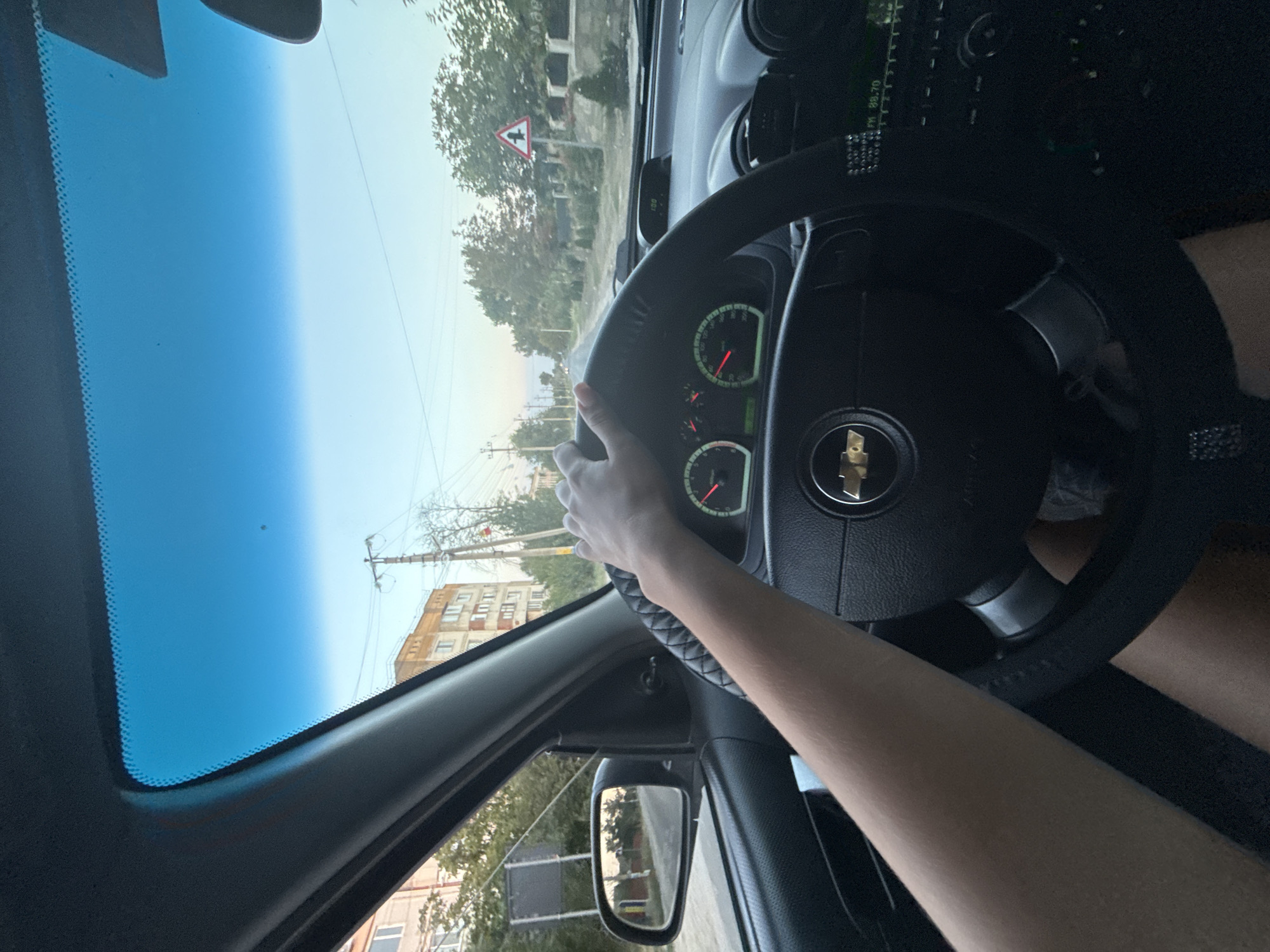 In Abaclia, Moldova, a person is driving a car. The photo is taken from the perspective of the driver, showing their left arm and hand on the steering wheel. The car's dashboard, including the speedometer and tachometer, is visible. Through the windshield, a residential street can be seen, with buildings and trees lining the road. A traffic sign indicating a turn is visible in the distance. The sky is clear and blue.
