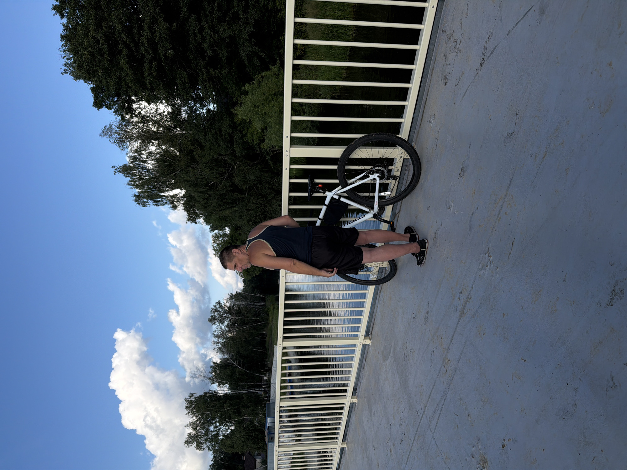 An adult male is standing on a paved bridge, looking to the left. He is wearing a dark tank top, black shorts, and black sneakers. Beside him, leaning against the bridge railing, is a white bicycle with black tires and a black seat. The bridge has a white metal railing with vertical bars. The background shows a body of water, dense green trees, and a blue sky with white clouds. The location is identified as Malakhovka, Russia.
