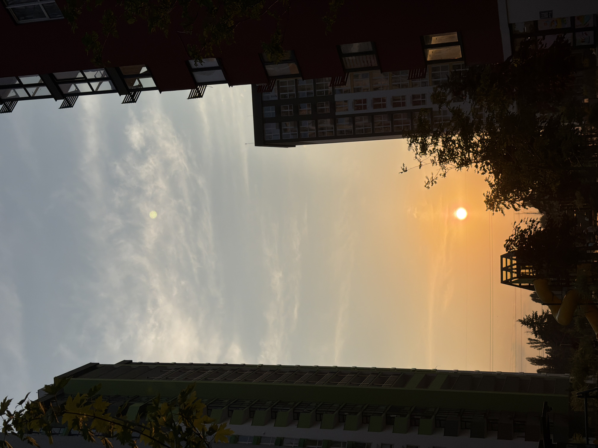 The image shows a view of apartment buildings in Kyiv, Ukraine, during sunset. In the foreground on the left is a tall, modern apartment building with a green facade and balconies. On the right is another apartment building with a dark red facade and visible windows. In the middle distance, a slightly shorter apartment building with a patterned facade is visible. The sky is filled with a gradient of colors, from pale yellow and orange near the horizon to a dusty blue at the top. The sun, a bright orb, is visible near the horizon. Silhouetted trees and a playground structure are present in the lower part of the image, suggesting an urban residential area. The scene is captured from a low angle, looking up towards the sky.