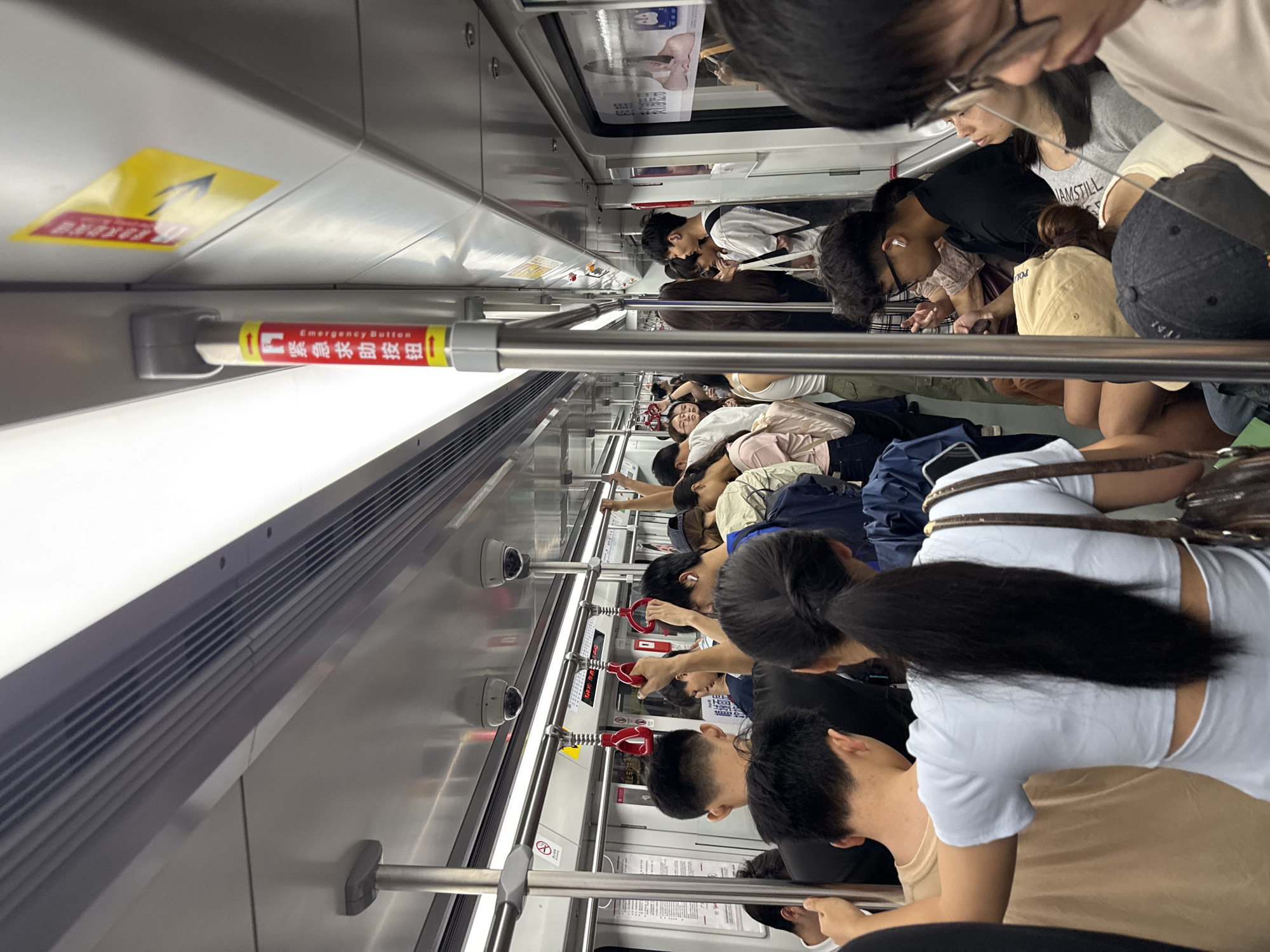 In Haizhu District, China, a crowded subway car is depicted, filled with passengers standing and seated. The interior of the subway is characterized by metal poles for support, ceiling-mounted security cameras, and overhead luggage racks. Red emergency handles are visible, as well as a red and yellow sign on a pole with Chinese characters and the English text "Emergency Button." A yellow sign with Chinese characters is also affixed to the ceiling. Passengers are dressed in casual clothing, with some looking at their phones. The scene captures the typical rush hour commute within a metropolitan public transportation system.