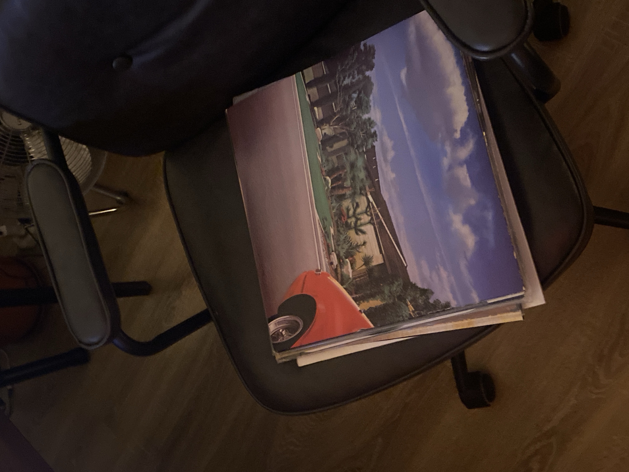 A stack of vinyl records rests on the seat of a black office chair. The album covers show landscapes with buildings and a sky. A partial view of a fan and a basketball are visible to the left. The scene is indoors, with a wooden floor. The location in the city of Шебунино, Russia is not discernible from the image.