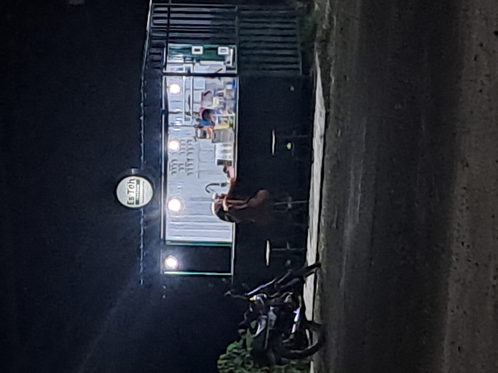 The image depicts a night scene in Slambur, Indonesia, featuring a small roadside food stall named "Es Teh." The stall is illuminated by interior lights, showcasing a counter with various containers and what appears to be a beverage dispenser. A person is seated at the counter, leaning forward, their back to the camera, wearing a dark shirt and a reddish-brown backpack or jacket. In front of the stall, a motorcycle is parked on the side of a dirt or asphalt road. The surrounding environment is dark, with some greenery visible to the left of the stall. The overall impression is of a simple, local establishment operating after dark.