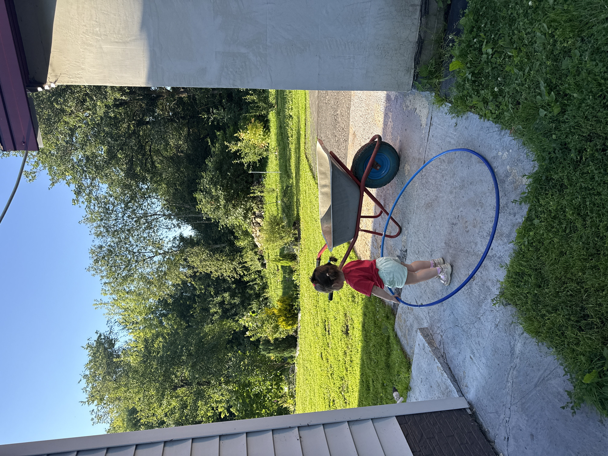 A young child, seen from behind, stands on a concrete patio near a white-sided building in Bolshie Ozerski, Russia. The child, wearing a red t-shirt and light-colored shorts, holds a blue hoop. A red and blue wheelbarrow is situated to the right of the child, positioned on the patio. The background features a lush green lawn, a line of trees, and a stone wall. The sky is clear and blue.