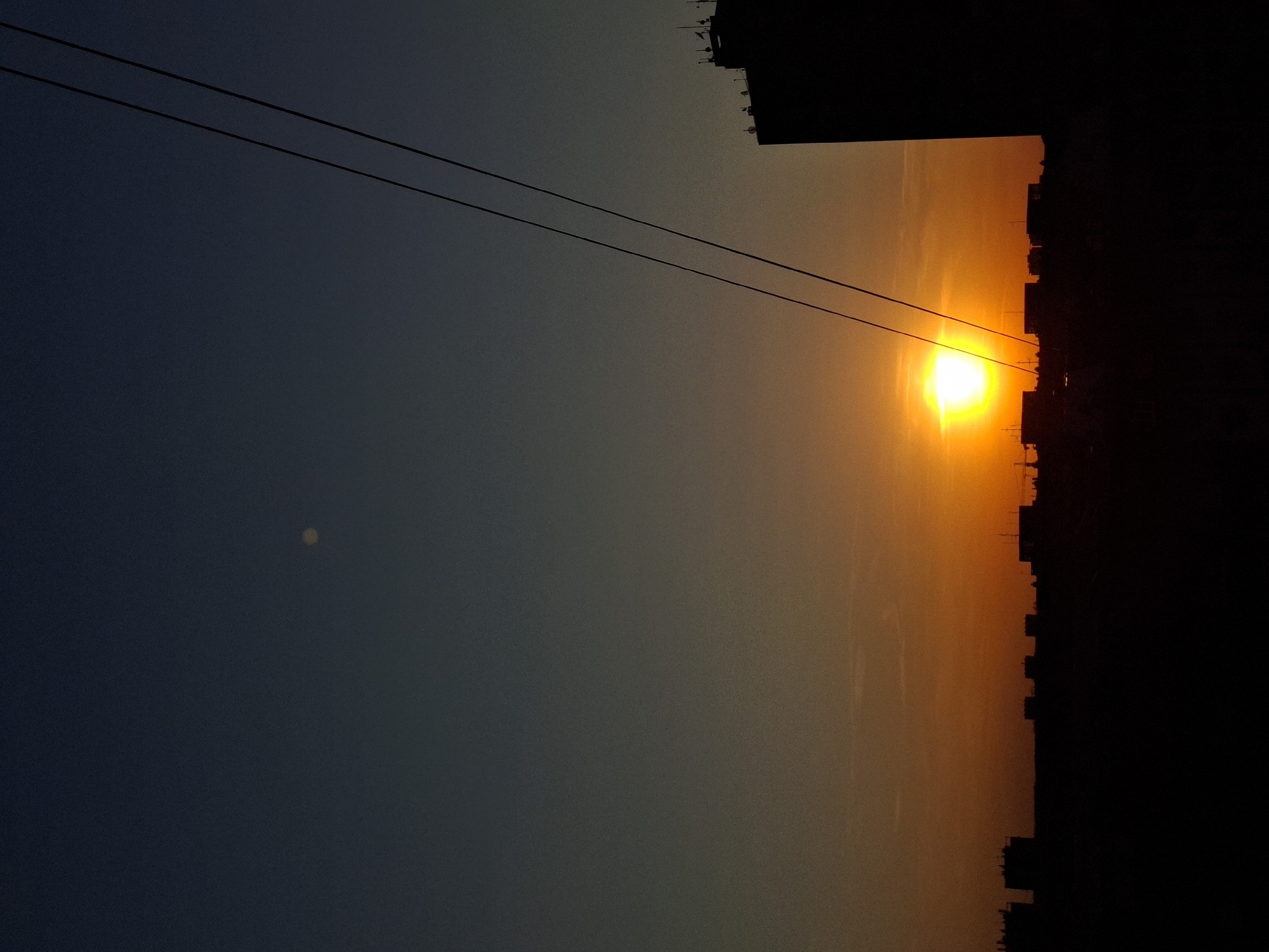 The image captures a vibrant sunset or sunrise over an urban landscape in Novi Sad, Serbia. The sky displays a dramatic gradient of colors, transitioning from a dark blue-grey on the left to a warm, hazy orange and brilliant yellow on the right. A prominent, intensely bright and glowing orb, likely the sun, is positioned low on the horizon on the right side, casting a strong golden light across the clouds and atmosphere.
Silhouetted against this colorful sky are the dark, jagged forms of buildings and rooftops, forming an urban skyline along the bottom edge of the frame. More building structures are visible in silhouette along the top right corner. Two dark, thin power lines or cables stretch horizontally across the mid-ground of the sky, creating a linear element that divides the expansive view. In the darker, upper-left portion of the sky, a faint, circular yellowish spot, possibly a lens flare or dust particle on the lens, is visible.
The scene depicts the natural event of a sunset or sunrise, with the city's architecture forming a striking contrast against the vivid sky. The time of day is clearly either early morning or late evening, given the low position of the sun and the long shadows or silhouettes. The overall impression is one of serene beauty during a transitional moment of the day. There is no visible text in the image.