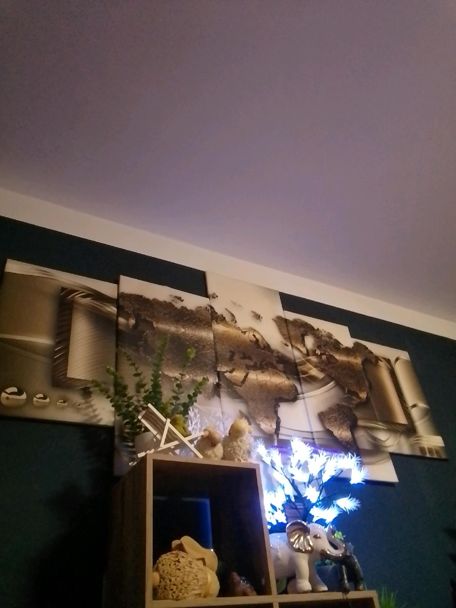 The image shows a close-up, low-angle shot of a wall decoration in a room. The main focus is a large, multi-panel artwork depicting a stylized world map with a textured, weathered appearance. To the left of the map, on the wall, are three spherical decorative objects. Below the artwork, a wooden shelf unit is visible. On the top shelf of the unit, there is a small green plant in a pot, a miniature white deck chair, and two small decorative figures, possibly birds or sheep. Also on the top shelf, to the right, is a decorative white tree with blue lights. On the shelf below, a wicker-like object, possibly a decorative basket or figure, is visible. A small white elephant figurine stands on the shelf to the right of the wicker object, next to a smaller dark figurine, likely another animal. The room's walls are dark blue, and the ceiling is a light purple-gray. The image is taken indoors, likely in a residential setting. The presence of Christmas-like decorative lights on the tree suggests a festive season. There are no people visible in the image. There is no information to indicate this location is in the city of Wedel, Germany.