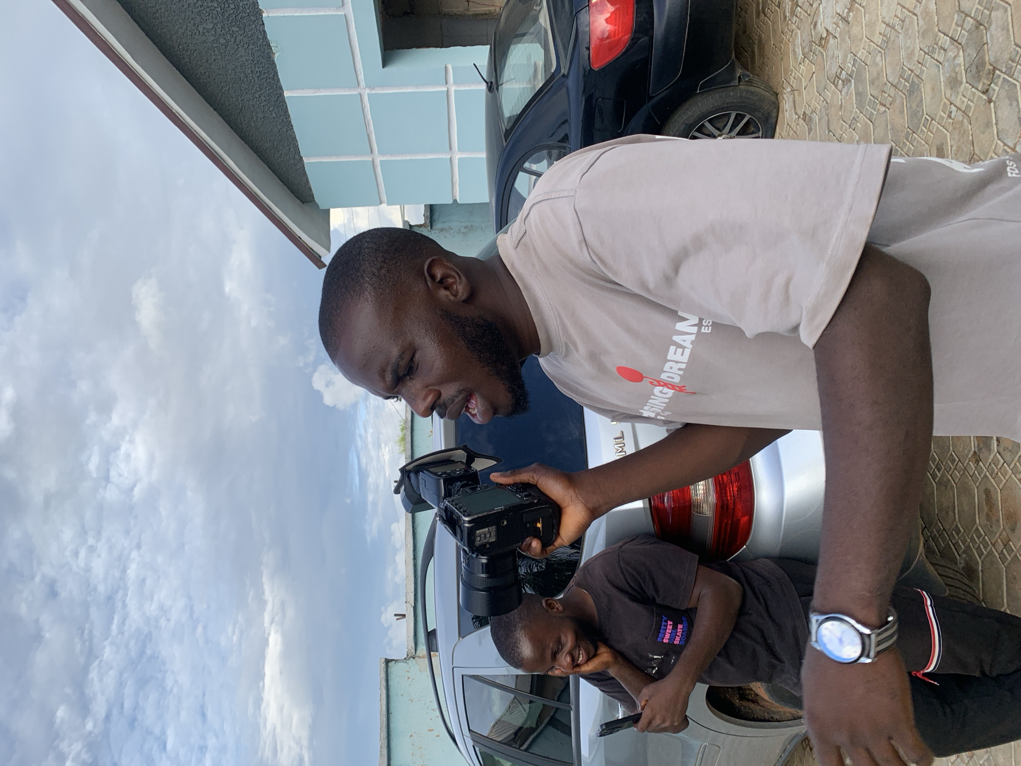 This image depicts an outdoor scene in the city of Jos, Nigeria, featuring two adult males and two vehicles against a backdrop of residential buildings and an overcast sky.
In the foreground, slightly to the right of the frame, a dark-complexioned man with short dark hair and a short beard is positioned. He is wearing a light tan short-sleeved t-shirt with a red circular graphic and white text that reads "RISING DREAM ES" on the front. His right arm is extended, holding a black professional-grade digital camera with a large lens attached. A small accessory is mounted on top of the camera. His head is turned to the left, and his mouth is open, as if he is speaking loudly or giving an instruction. On his left wrist, he wears a silver-colored analog watch with a striped grey and white fabric strap. His left arm is bent, with his hand mostly out of frame.
To the left, in the midground, another dark-complexioned man with short dark hair and slight facial hair is leaning against the rear of a silver SUV. He is wearing a dark brown/black short-sleeved t-shirt with white text that reads "PRETTY SWEET SKATEBOARDS." He is also wearing black trousers or sweatpants with a distinct red and white horizontal stripe detail near the left ankle. He is smiling broadly with his eyes partially closed, his right hand covering his mouth and chin, indicating hearty laughter. In his left hand, he holds a dark-colored smartphone vertically.
The silver SUV he is leaning on appears to be a Mercedes-Benz ML-Class, with its rear taillight and part of the bumper visible. Further into the background, behind the silver SUV, a black sedan is partially visible on the right, showing its rear quarter panel and taillight.
The ground is paved with light brown interlocking blocks arranged in a geometric pattern. In the background, on the right side of the image, stands a building with a dark brown sloped roofline, a light grey stucco-like upper wall, and a lower section clad in light blue square tiles, forming a grid pattern. To the far left in the background, another light blue building with a textured wall is partially visible. The sky above is overcast with scattered light grey and white clouds, suggesting a bright but diffused daylight condition. The overall scene conveys an informal, candid outdoor gathering or production setting.