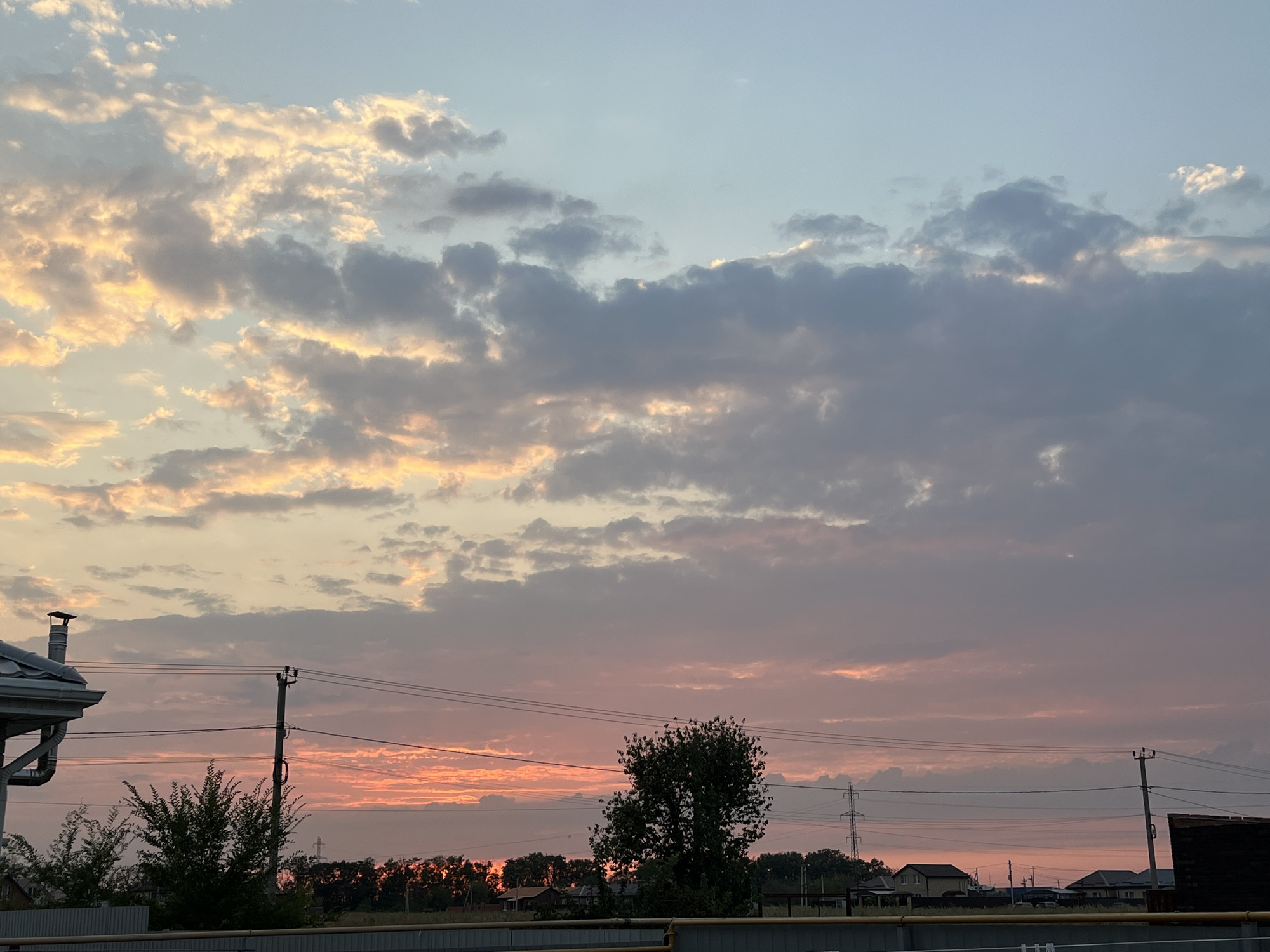 The image depicts a sunset scene over a suburban area in Taganrog City District, Russia. The sky is filled with a mix of orange, pink, and blue hues, with scattered clouds. In the foreground, a house with a gray roof and a chimney is visible on the left. Power lines and utility poles crisscross the frame, connecting to structures in the distance. A few trees are scattered throughout the scene, including a large, leafy tree in the middle ground. Several houses with varying architectural styles are visible in the background, interspersed with trees and vegetation. The overall impression is of a quiet, rural landscape during twilight.