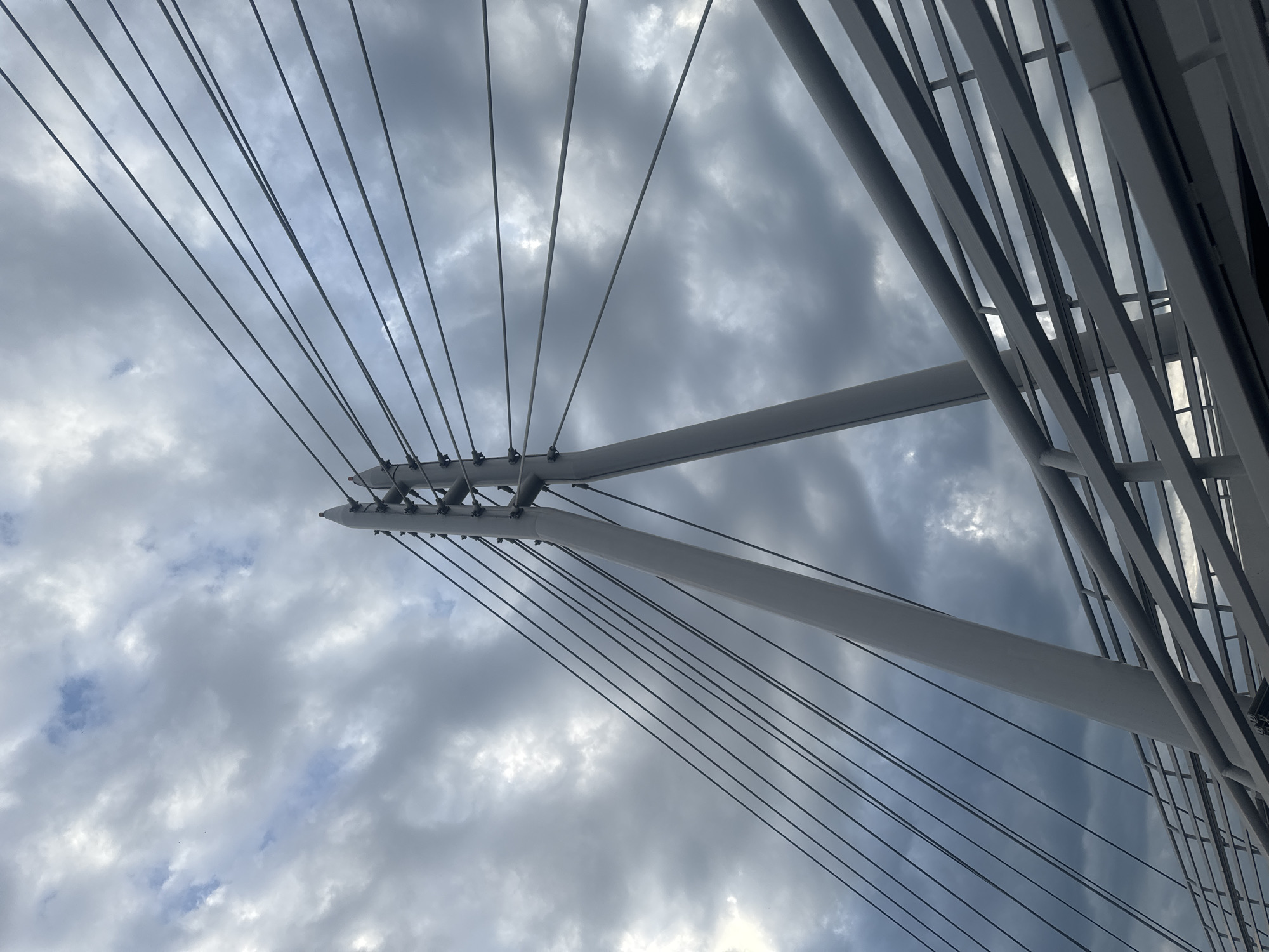 The image shows a low-angle view of a white cable-stayed bridge tower against a cloudy sky. Numerous thin cables extend from the tower, fanning out diagonally across the frame. A section of the bridge's railing, also white and composed of horizontal bars, is visible in the lower right quadrant. The sky is filled with dramatic, dark gray clouds interspersed with patches of lighter gray and a hint of blue. The overall impression is one of architectural strength and an overcast, potentially stormy, atmosphere. The image does not contain any people or specific landmarks that would identify it as being in Irkutsk, Russia.