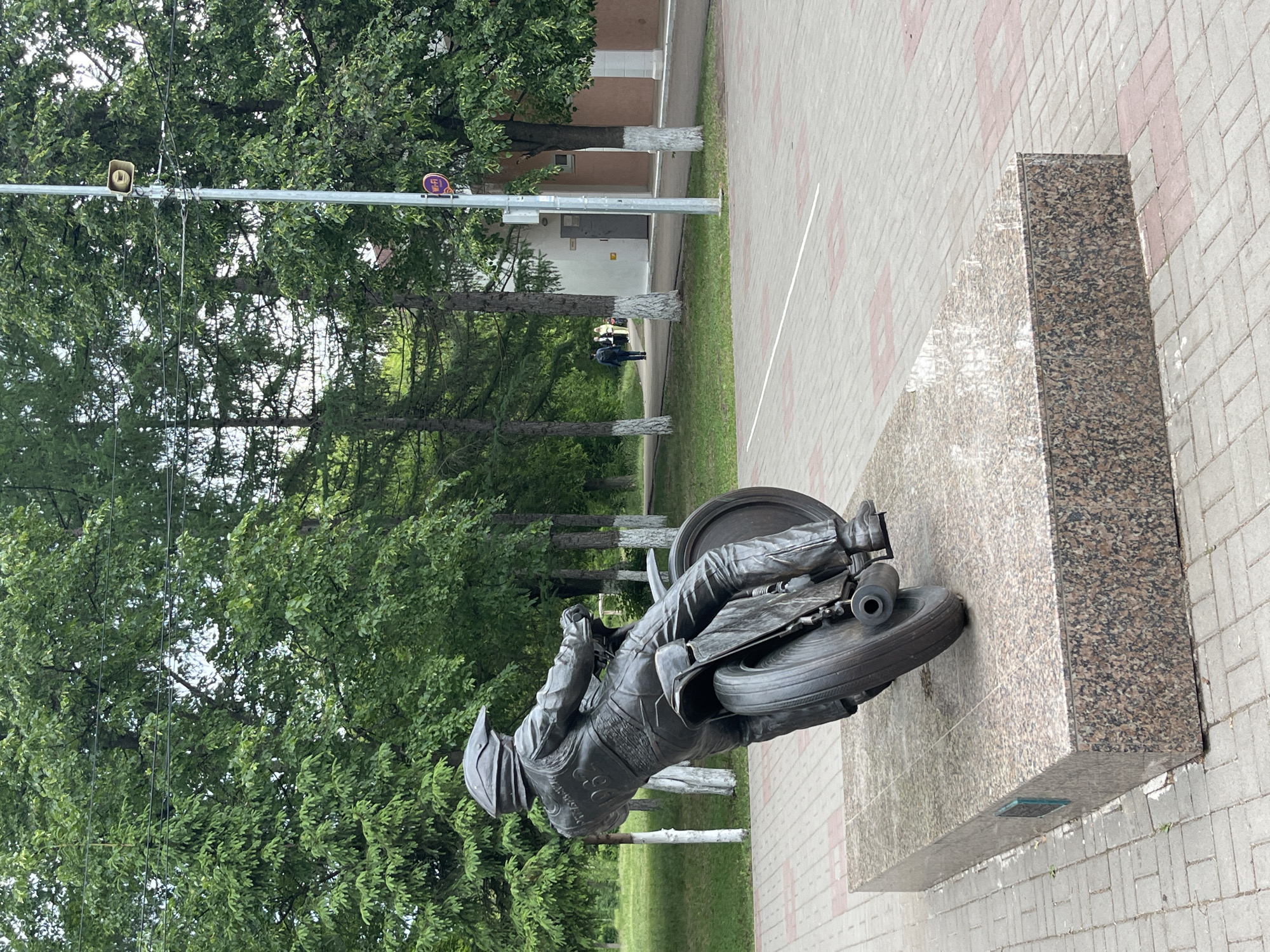 A static, daytime outdoor scene in Oktyabrsky, Russia, features a prominent bronze statue of a motorcyclist and motorcycle. The statue depicts a racer in a helmet and full gear, leaning sharply as if navigating a turn. The motorcycle appears to be a speedway or racing model. This monument rests on a large, polished granite pedestal with varied brown and black speckles, located on a paved plaza. The paving consists of interlocking blocks in shades of grey and reddish-brown.
Surrounding the plaza are numerous green trees, some with their lower trunks painted white, a common practice in the region. In the mid-ground, a light pole stands tall, equipped with electrical wires, a speaker-like device, and a circular purple sign. The sign displays the text "Wi-Fi" in white letters. Further in the background, a light-colored building with a dark door is visible, and two indistinct figures can be seen walking on a path nearby. The lighting suggests an overcast or bright daytime, with no harsh shadows and clear visibility. There is an unclear inscription on the statue's chest, possibly a racing number.
