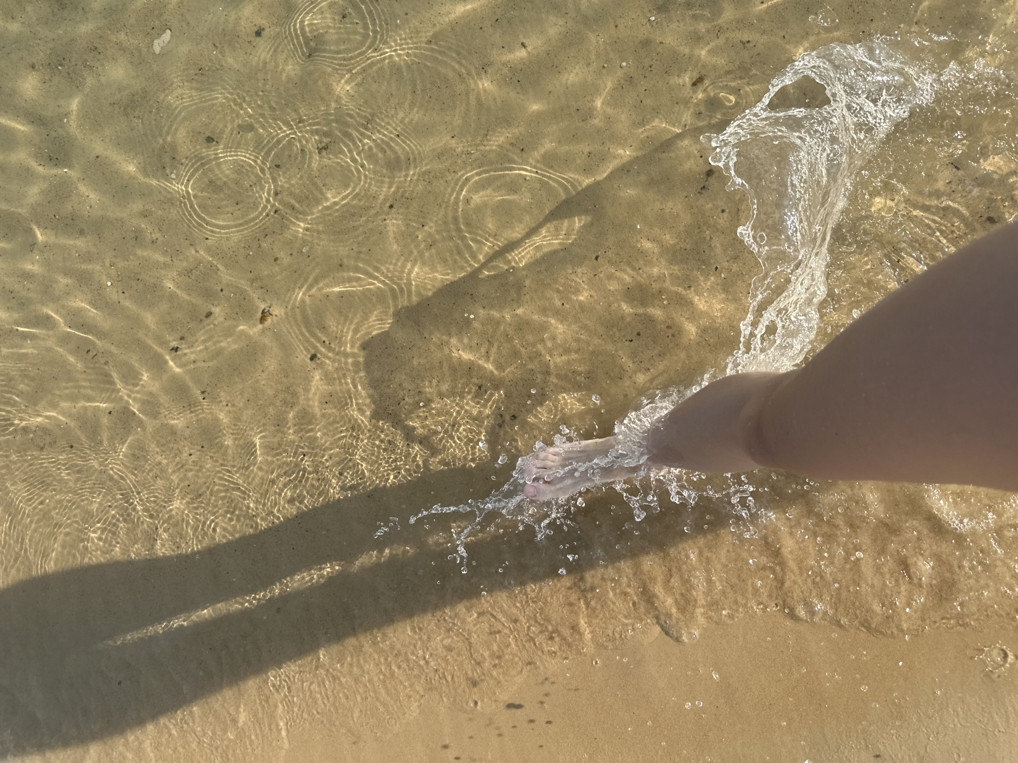 An overhead shot captures a person's bare leg and foot stepping into clear, shallow water over a sandy seabed. The foot is in the process of kicking up a significant splash of water, creating dynamic white foam and droplets. Long, dark shadows, likely cast by the person and potentially other unseen objects, stretch across the sandy bottom from the left side of the frame. Sunlight filters through the water, creating mesmerizing ripple patterns and bright, circular light reflections on the sand. The sand itself is light brown with small dark specks. The water is so transparent that the texture of the sand and small debris on the bottom are clearly visible. The scene suggests a sunny day, possibly morning or late afternoon given the elongated shadows, at a beach, lake, or calm body of water. No text is visible in the image.