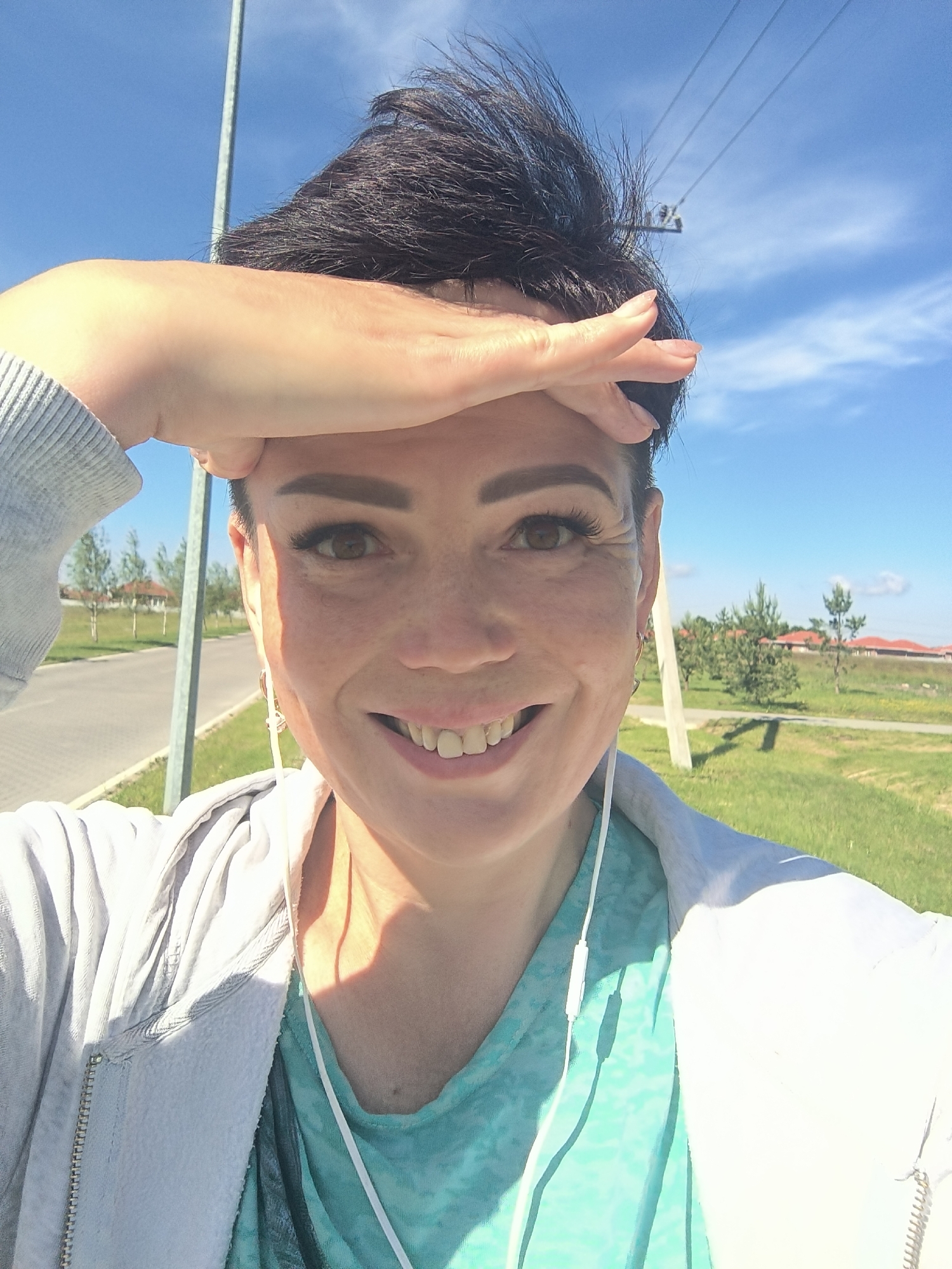In an outdoor selfie taken on a sunny day in Laskino, Russia, a woman with short, dark, spiky hair and fair skin smiles broadly at the camera. She is wearing a light grey zippered hoodie over a teal patterned t-shirt, and white earbuds are visible. Her right hand is raised to her forehead, shielding her eyes from the sun. The background shows a clear blue sky with wispy clouds, some trees and green grass, a paved road, and buildings with red roofs in the distance. A grey pole is visible to her left.