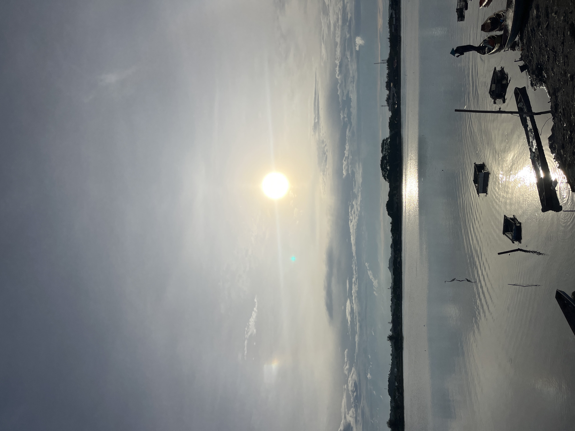 The image shows a waterfront scene in Lhokseumawe, Indonesia, at dawn or dusk. The sun is visible through a cloudy sky, reflecting on the still water. Several small boats are moored near the shore, some appearing damaged or partially submerged. The shoreline consists of dark earth or mud. The opposite shore has trees. In the distant background, there are towers.