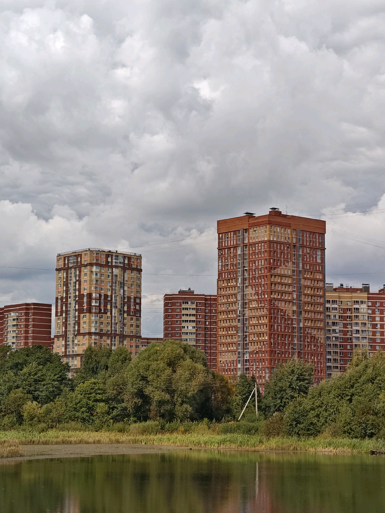 The image depicts a residential area in Shcherbinka, Russia, with a focus on several multi-story apartment buildings. The buildings are constructed with a combination of red brick and beige panels, featuring numerous windows and balconies. In the foreground, a body of water reflects the sky and the surrounding greenery. Tall trees and vegetation line the edge of the water, separating it from the buildings. The sky is overcast with dense, grey clouds, suggesting a cloudy day. Utility poles and wires are visible in the background, indicating urban infrastructure. The scene appears to be a typical view of a modern housing development in a suburban or semi-urban environment. There are no people visible in the image.