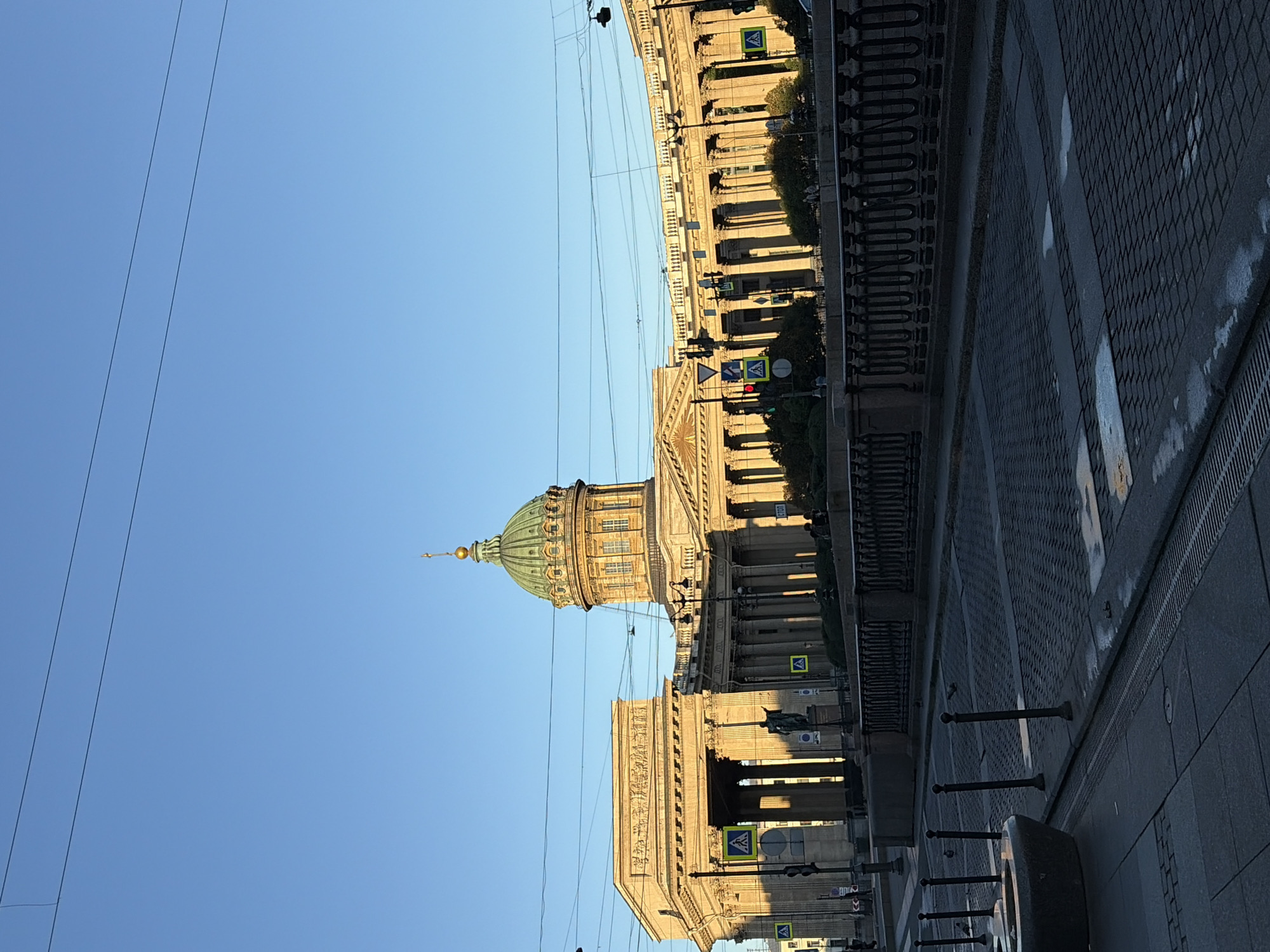 This is a wide-angle, outdoor, eye-level photograph taken in Saint Petersburg, Russia. The image captures the Kazan Cathedral, a prominent landmark, with its distinctive semicircular colonnade stretching out to the sides. The cathedral features a large, green dome topped with a golden spire, set against a clear, bright blue sky. Sunlight casts long shadows from the architectural elements, indicating it might be late afternoon or early morning.
In the foreground, a cobblestone street leads towards the cathedral. A black, ornate metal railing runs along what appears to be a canal or riverbank in front of the building. Several dark, slender bollards are spaced along the sidewalk. A stone-like object, possibly a planter or fountain base, is visible on the left side of the street. Pedestrian crossings are marked with yellow, diamond-shaped signs featuring a pedestrian symbol. Traffic lights are also present. A few people are visible in the background, appearing as small figures near the colonnade and on the sidewalk. Overhead power lines are visible crisscrossing the sky. The overall impression is of a calm, historic urban scene bathed in warm light.