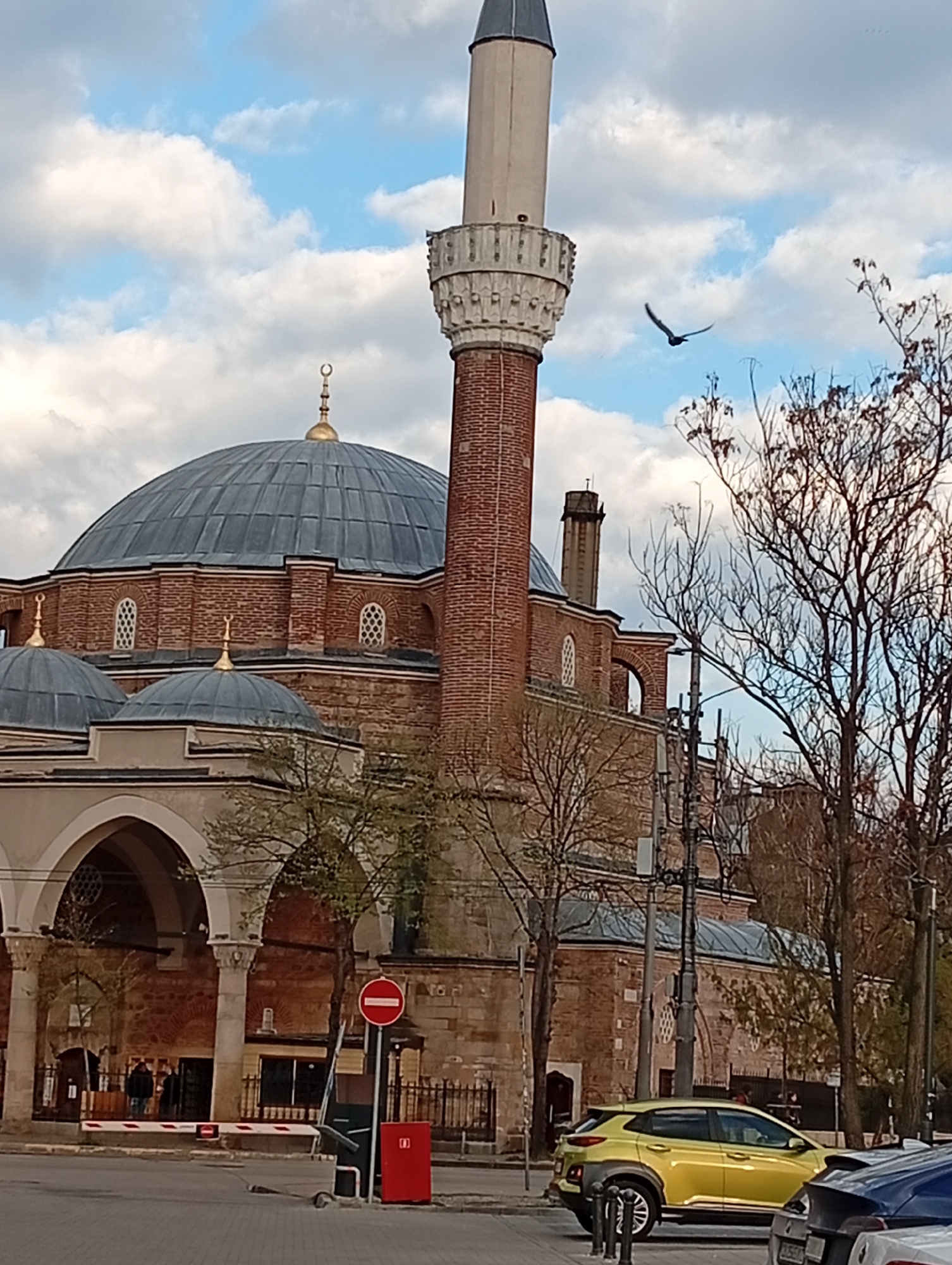 The image depicts the Banya Bashi Mosque in Sofia, Bulgaria, under a partly cloudy sky. The mosque's distinctive minaret and large dome are prominent. A bird is in flight near the minaret. In the foreground, there is a yellow Hyundai Kona parked on the street. A red mailbox and a "Do Not Enter" sign stand on the sidewalk. Bare trees are visible on the right side of the image. Several people can be seen near the mosque's entrance. The image appears to be taken at street level.