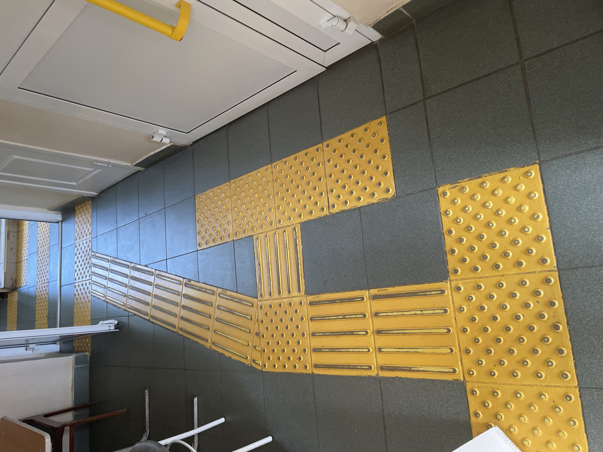 The image shows a section of a floor with tactile paving. The tiles are yellow and have raised patterns to aid navigation for people with visual impairments. Some tiles have small bumps, while others have raised lines. The tiles are laid on a gray tiled floor and lead towards a closed white door or hatch on the ceiling. The scene appears to be indoors, possibly a public space or transportation terminal, given the presence of the tactile paving. There are legs of a chair and table in view. There is no specific location within Chaikovsky, Russia visible in this image.