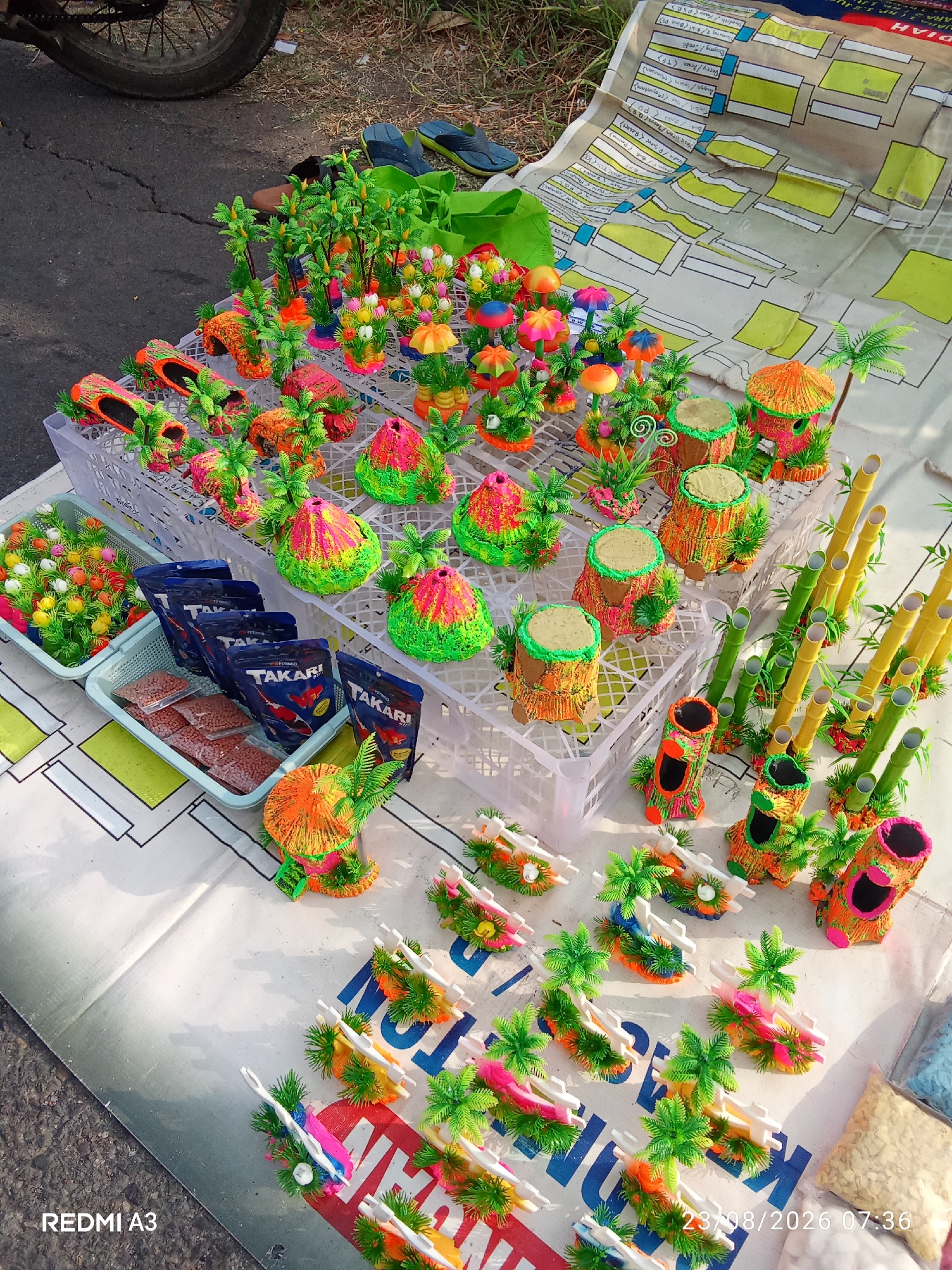 The image depicts a street stall in Grogol, Indonesia, displaying a variety of colorful, artificial decorations likely intended for aquariums or gardens. In the foreground, numerous small, vibrant ornaments are laid out on a white cloth patterned with a grid and some text. These include palm tree-like structures, whimsical mushroom shapes, and volcano-shaped pieces, all adorned with bright pink, orange, green, and yellow hues.
Behind these small decorations, larger items are arranged on a white plastic crate. These are more elaborate versions of the smaller ornaments, featuring detailed palm trees, houses resembling thatched huts, and hollow logs, all similarly decorated in vivid colors. To the left of the crate, two pale blue plastic baskets are visible. One contains a colorful arrangement of small, artificial flowers. The other basket holds several sealed plastic bags of what appears to be fish food, with the "TAKARI" brand clearly visible on some of them.
To the right of the crate, several stalks of artificial bamboo in green and yellow are arranged vertically. Further to the right, more of the colorful, hollow log-shaped ornaments are displayed. In the lower right corner, several bags of what appear to be small stones or gravel are visible.
The background of the image shows a darker asphalt surface, likely a road or sidewalk, with a portion of a bicycle wheel on the left. Some grass and greenery are also visible in the upper portion of the image, suggesting an outdoor setting. A bright green bag and a pair of blue flip-flops are also present in the background, hinting at the presence of the stall owner or other people. The overall scene is one of a lively street market display, emphasizing the vibrant and artificial nature of the merchandise.