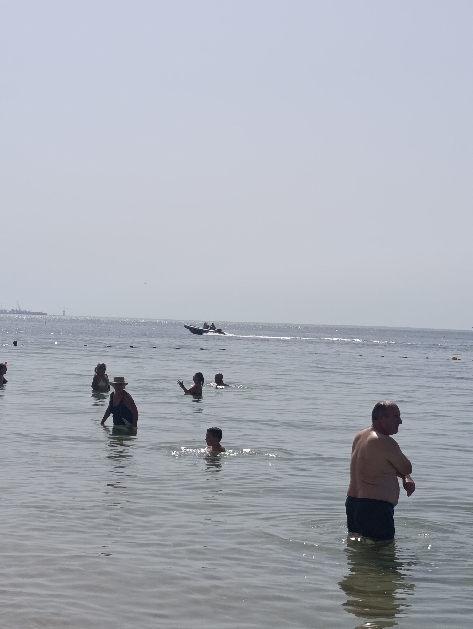 An overhead, wide shot captures an outdoor scene on a hazy, bright day, likely at a beach in Setúbal, Portugal. The foreground is dominated by the calm, shallow water of the sea, with gentle ripples reflecting the light. Several people are partially submerged in the water, engaged in various activities. In the foreground, a middle-aged man with a light tan torso and dark swim trunks is visible from the waist up, facing away from the camera, his arms bent at the elbows. Behind him and to his left, a child with dark hair is also in the water, facing left. Further back and to the left, a woman wearing a dark swimsuit and a wide-brimmed hat stands facing the camera. To her left, another woman in a white bikini is visible, facing left. Several other individuals are dispersed in the water, some fully submerged and others waist-deep. In the mid-ground, a motorboat with two occupants speeds across the water, leaving a white wake. The background consists of the expansive, hazy sea, meeting a pale, almost white sky. In the far distance, faint outlines of industrial structures and a large ship suggest a port or industrial area on the horizon. The overall impression is one of leisure and recreation on a warm, sunny day at the coast.