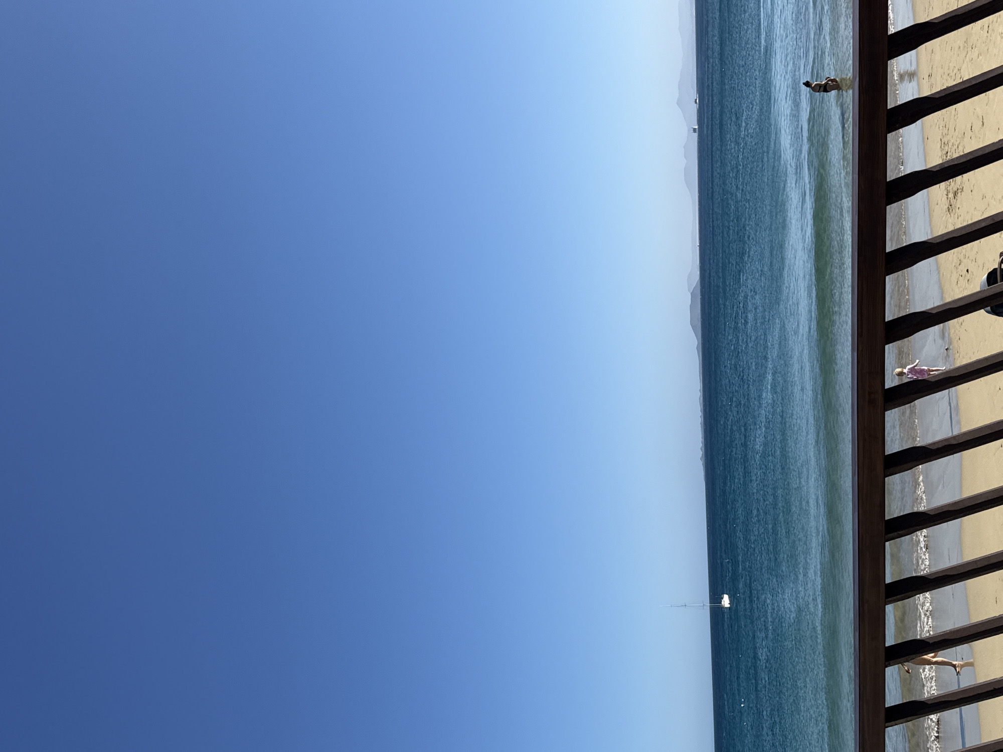The image depicts a sunny day at Playa Blanca, Spain. The right side of the view is partially obscured by a wooden railing. Beyond the railing, a sandy beach meets the blue ocean. Several people are on the beach and in the water. A sailboat is visible in the distance. The sky is clear and blue.