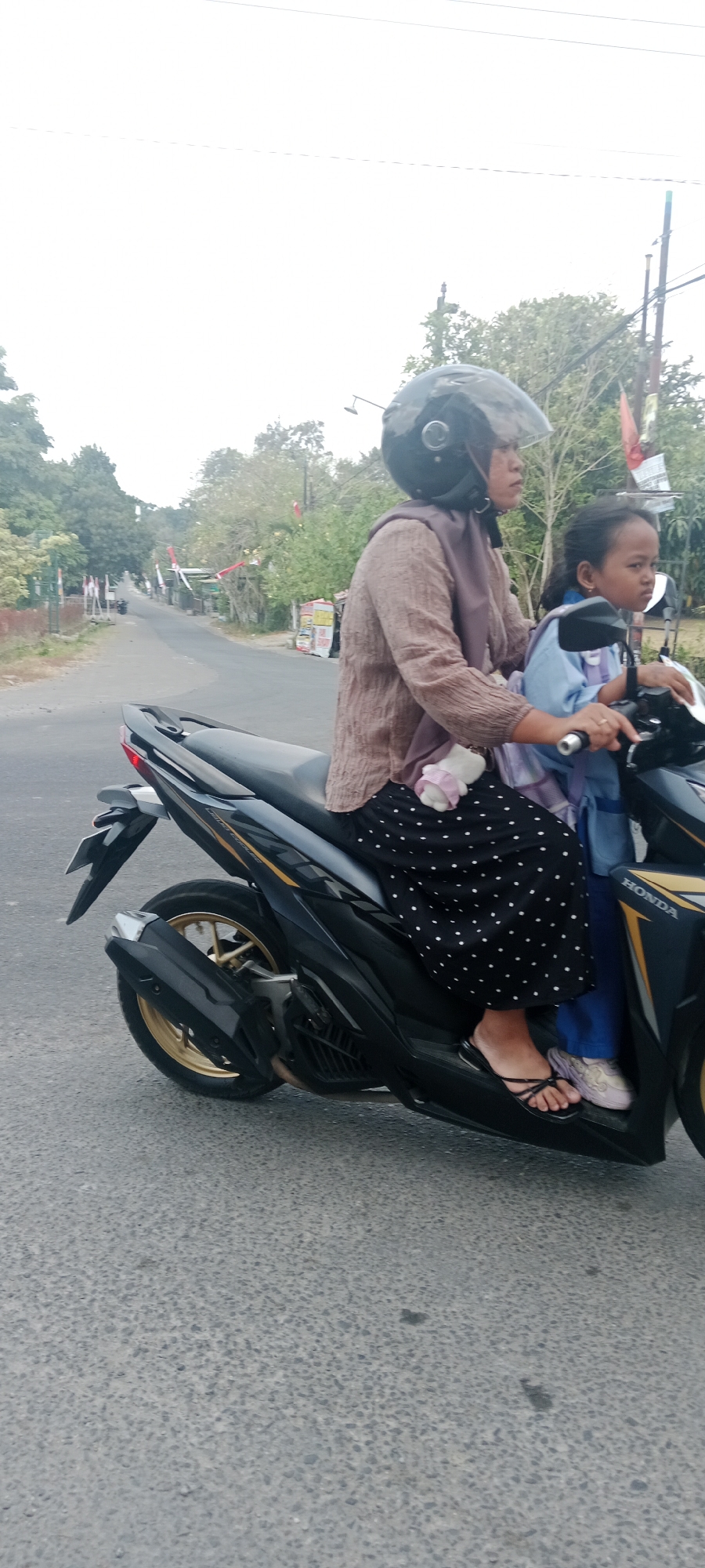 In Maguwoharjo, Indonesia, two individuals, an adult and a child, are riding a black Honda motorcycle on an asphalt road. The adult, wearing a light brown long-sleeved shirt and a headscarf, is driving the motorcycle. They are also wearing a black helmet with a clear visor. The child, seated in front of the adult, is wearing a light blue long-sleeved garment. Both individuals are looking forward. The motorcycle has a black body with gold accents on the wheels and exhaust pipe. The background shows a street lined with trees and some buildings, with red and white flags visible along the roadside, suggesting a festive or national event. The sky is overcast.