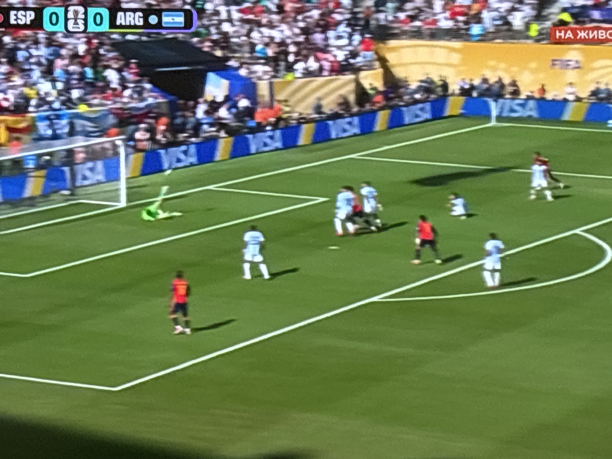 The image depicts a football match being played in a stadium. In the foreground, several players in red and light blue kits are visible on the green grass pitch. One player in a red kit is standing near the right corner of the penalty box, facing away from the camera. Another player in a light blue kit is positioned in the center of the frame, also facing away. In the background, more players can be seen, as well as a goalkeeper in a green kit who appears to have fallen to the ground near the goal on the left side of the image. A large crowd of spectators fills the stands behind the pitch. A scoreboard at the top of the image indicates the score as ESP 0, ARG 0. Advertising boards, including ones with the VISA logo, are visible along the side of the pitch. The stadium appears to be hosting a professional football game. The image does not contain any information that would allow for the identification of the city of Burgas, Bulgaria, or any specific location within it.