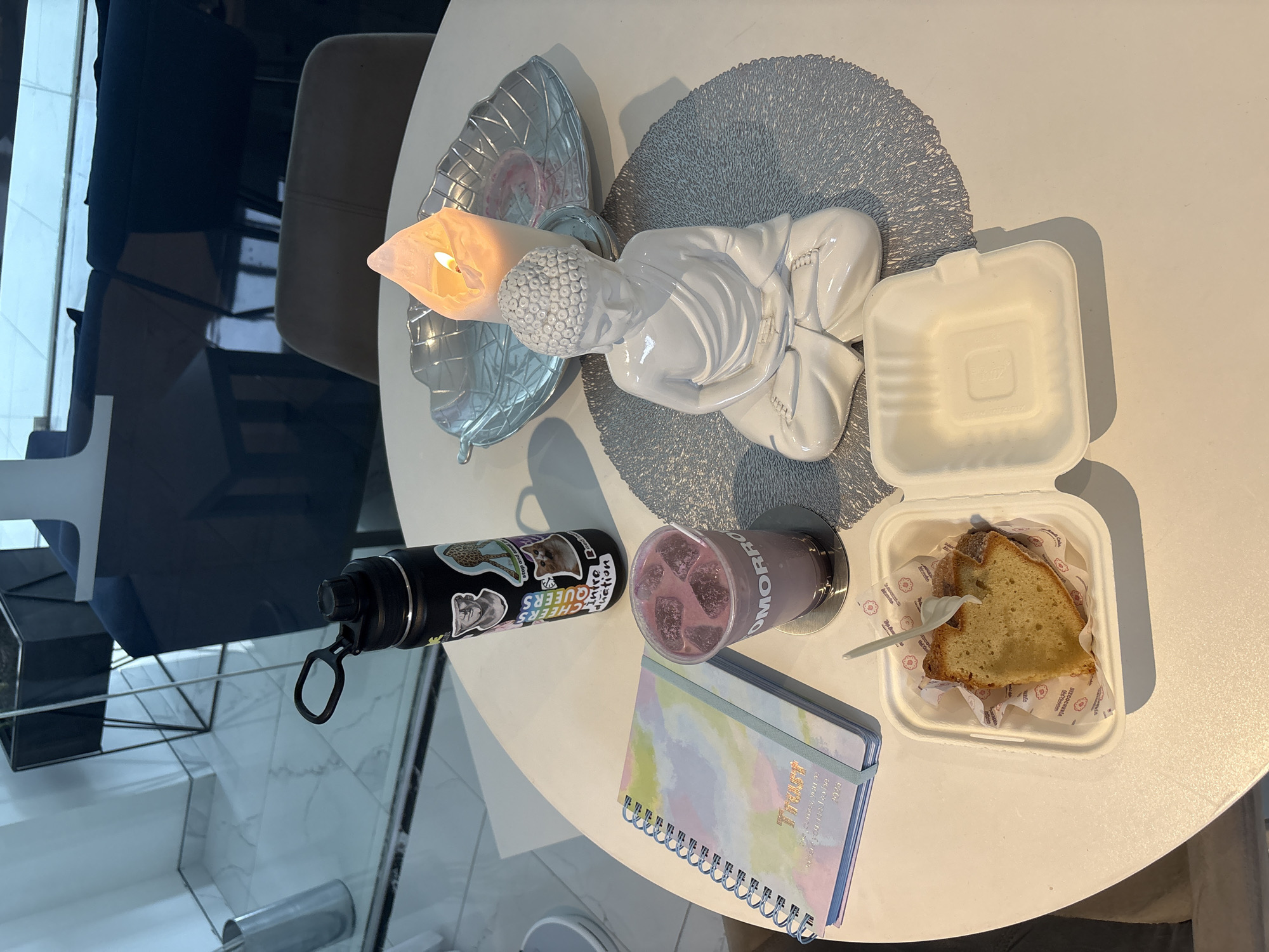 The image captures a table setting in what appears to be a cafe or restaurant, possibly located in Puebla, Mexico, judging by the brand of the drink.
On the white circular table, there's an assortment of items, suggesting a moment of relaxation or a break. A decorative candle holder with a lit candle sits alongside a white Buddha statue on a gray placemat.
Nearby is a partially open white takeout container, revealing a slice of cake. There's also a purple drink with ice, and a water bottle adorned with stickers. A notebook with the word "Trust" printed on it is also present. The environment appears to be well-lit, and the overall setting suggests a modern and comfortable space.