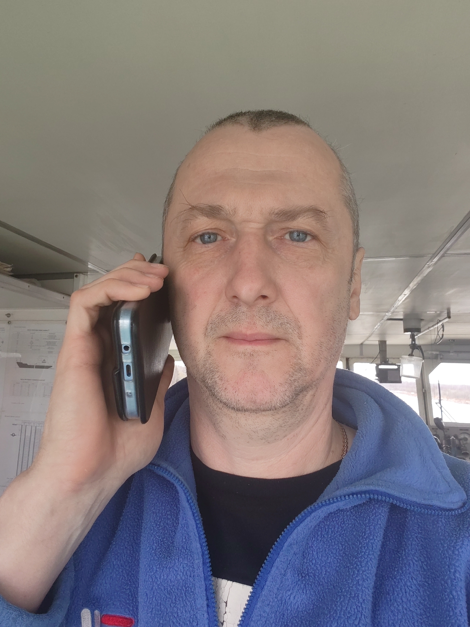 A middle-aged man with blue eyes and short, receding hair is holding a smartphone to his left ear, seemingly engaged in a phone conversation. He is wearing a blue fleece jacket over a black shirt, with a silver chain visible around his neck. The background suggests an interior space, possibly a control room or a cabin, with paneling and equipment visible. There are also papers posted on a wall. There is no evidence or indication to suggest that this image was taken in Uzhla, Russia.