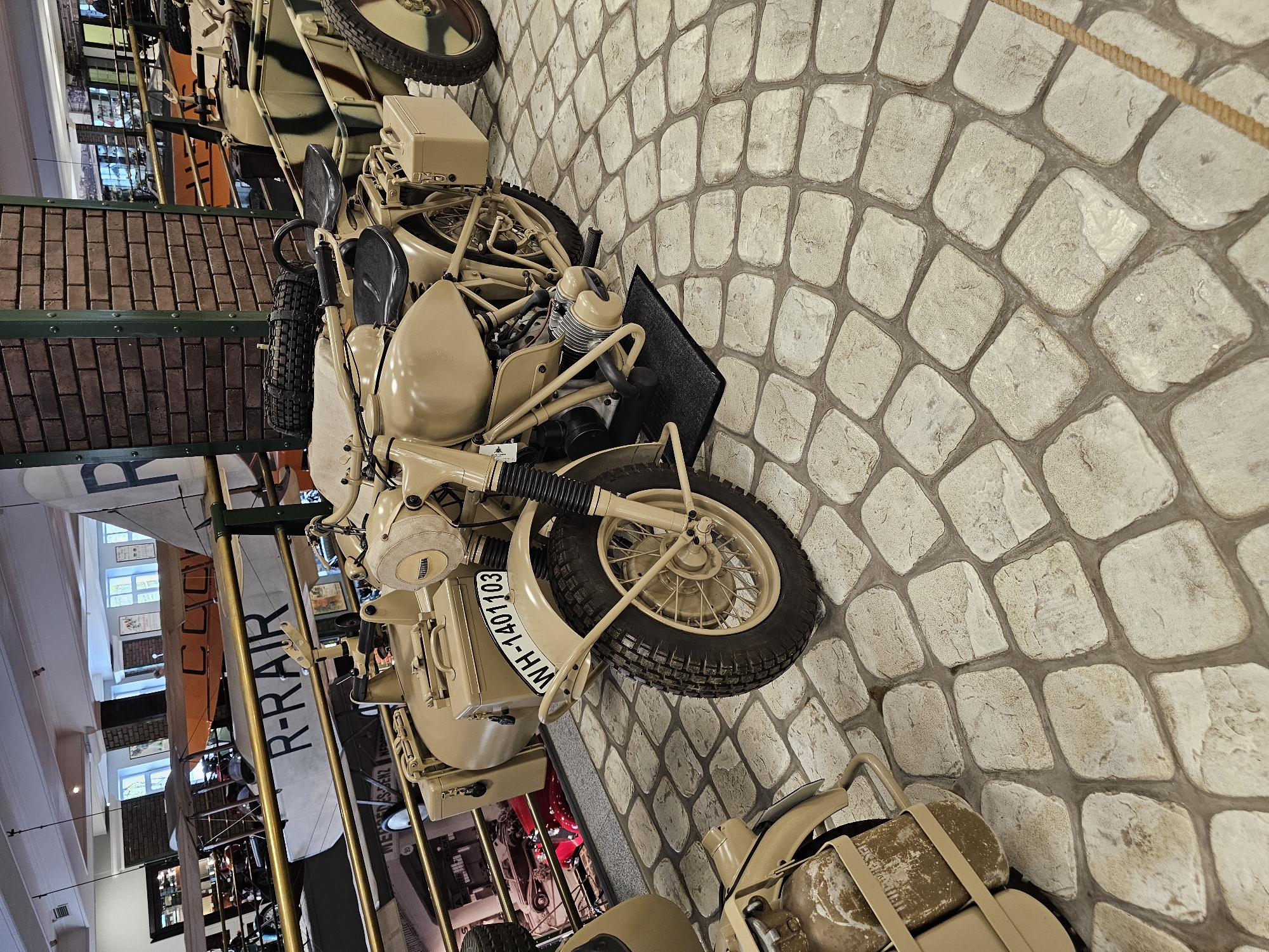 The image shows a vintage motorcycle with a sidecar, painted in a sand color. The motorcycle's license plate reads "WH-140103". The scene appears to be inside a museum, possibly in the city of Архангельское, Russia, given the request context. The motorcycle is on a stone-paved floor. In the background, elements of other exhibits are visible, including parts of a biplane labeled "R-RAR." The environment suggests an aviation and/or military history museum.