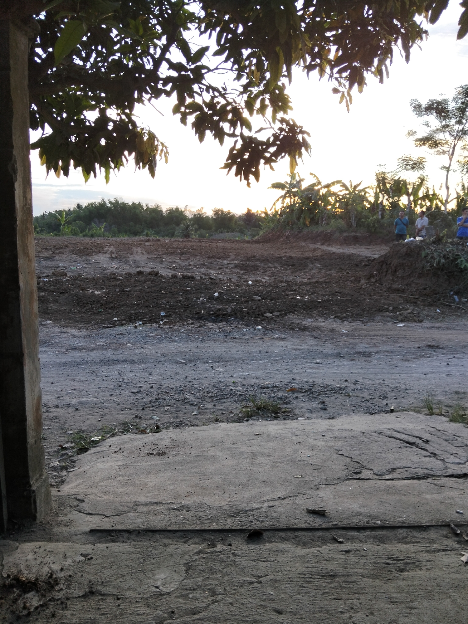 The image depicts an outdoor scene, likely in an undeveloped or construction area, with a focus on a patch of cleared, dark soil in the mid-ground. The sky is bright with a setting or rising sun, casting a warm glow and silhouetting some trees in the distance. In the foreground, a rough, concrete surface is visible, appearing somewhat cracked and weathered. To the left, a portion of a concrete pillar or wall is present, framing the view.
In the mid-ground, beyond the cleared soil, a line of dense green foliage, likely trees and vegetation, forms a backdrop. The cleared area itself shows signs of disturbance, with uneven mounds of earth and scattered debris, including small pieces of white and dark material. Towards the right side of the mid-ground, three individuals are standing on a mound of earth, facing away from the camera. The person on the far left wears a blue shirt, the middle person a light-colored shirt, and the person on the far right a blue shirt. They appear to be observing the area or engaged in a discussion.
The overall impression is of a transitional space, perhaps a site undergoing preparation for development, with natural elements meeting signs of human activity. The light conditions suggest it is either early morning or late evening. The location is specified as Payagaboh, Indonesia.