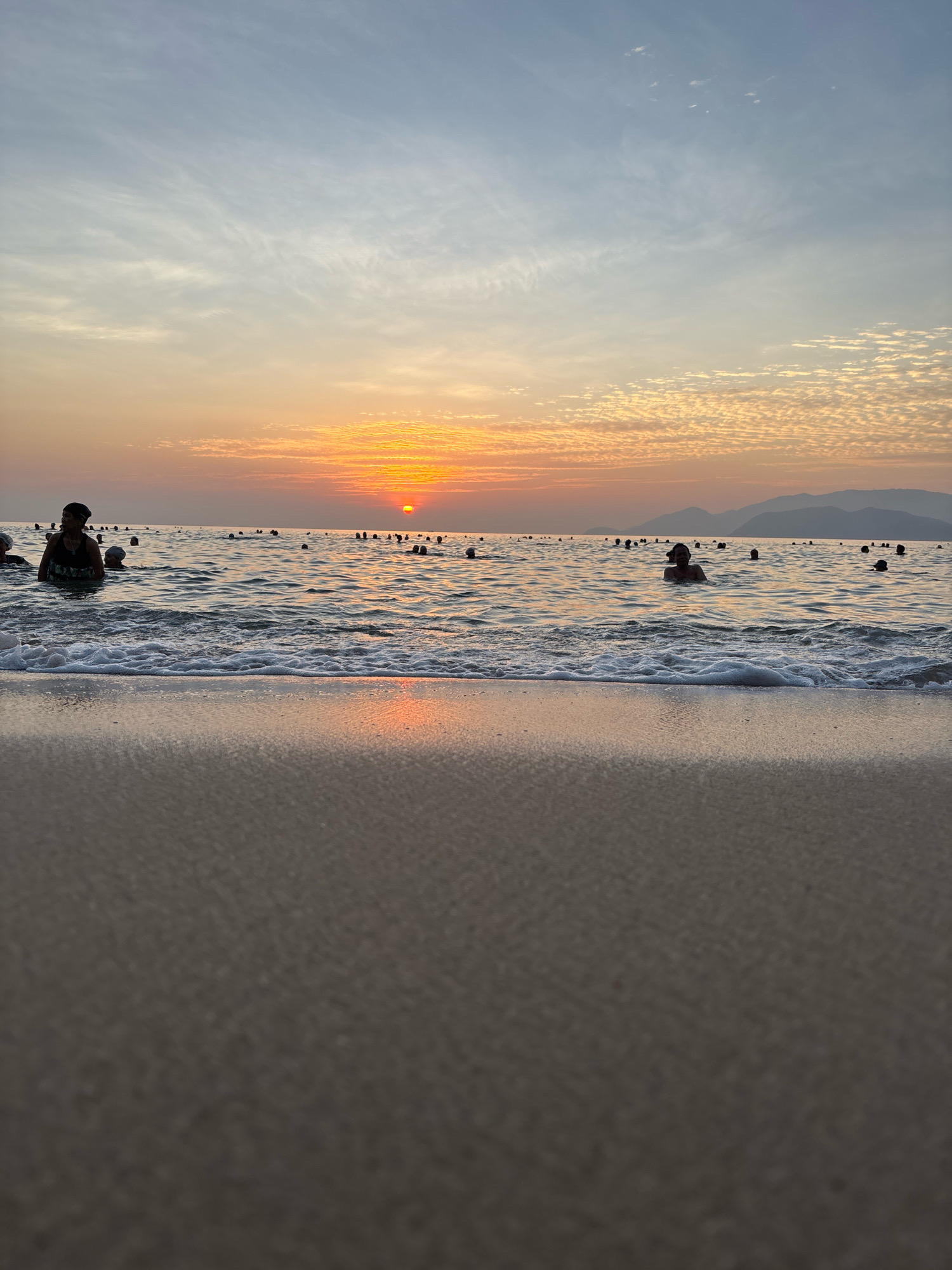 This is a photograph taken at a beach in Nha Trang, Vietnam, during sunset. The foreground is dominated by a sandy beach, with gentle waves lapping at the shore. In the middle ground, the ocean is populated by numerous people swimming and wading. The sun is setting on the horizon, casting a warm, orange glow across the sky and reflecting on the water's surface. The sky itself displays a gradient of colors, from a pale blue at the top to warmer oranges and pinks near the horizon, with scattered clouds. Distant mountains are visible in the background. The overall impression is one of a peaceful and lively beach scene at dusk.