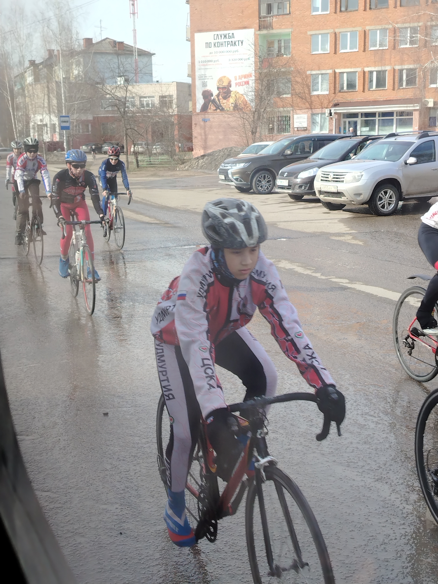 A group of young cyclists are riding on a wet asphalt road in Noviy, Russia. The cyclist in the foreground wears a white, red, and black jacket with "ЦСКА Удмуртия" written in Cyrillic on the sleeves and legs, a gray helmet, and blue and orange cycling shoes. Other cyclists, similarly dressed, follow behind. Cars are parked on the right side of the road. A brick building in the background has a large sign advertising contract service with the Russian army.