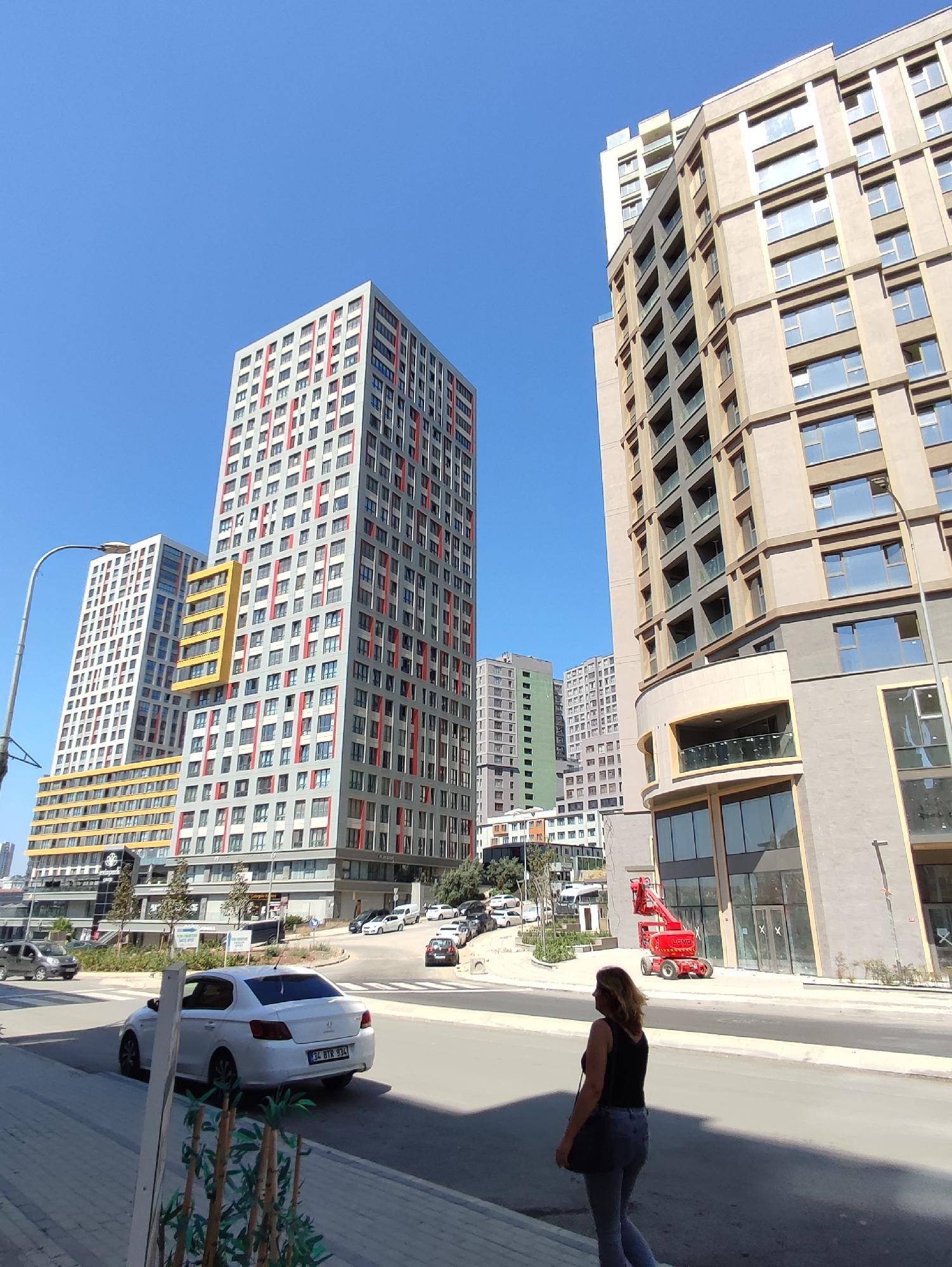 In Kadıköy, Turkey, a modern urban scene unfolds under a clear blue sky. A woman with blonde hair, wearing a black tank top and blue jeans, walks away from the camera on a paved sidewalk. Her dark shoulder bag is slung across her body. In the background, a bright red articulated boom lift is parked on the street, suggesting ongoing construction or maintenance. Across the street, a white sedan with license plate "34 BTR 934" is driving away. The street is lined with several contemporary high-rise apartment buildings, one predominantly light brown with numerous windows and balconies, and another taller building with a striking pattern of alternating grey sections and red and white vertical stripes. The architecture is modern and geometric, with many windows and balconies visible. The scene is characteristic of a developing urban area with a mix of residential and commercial structures.