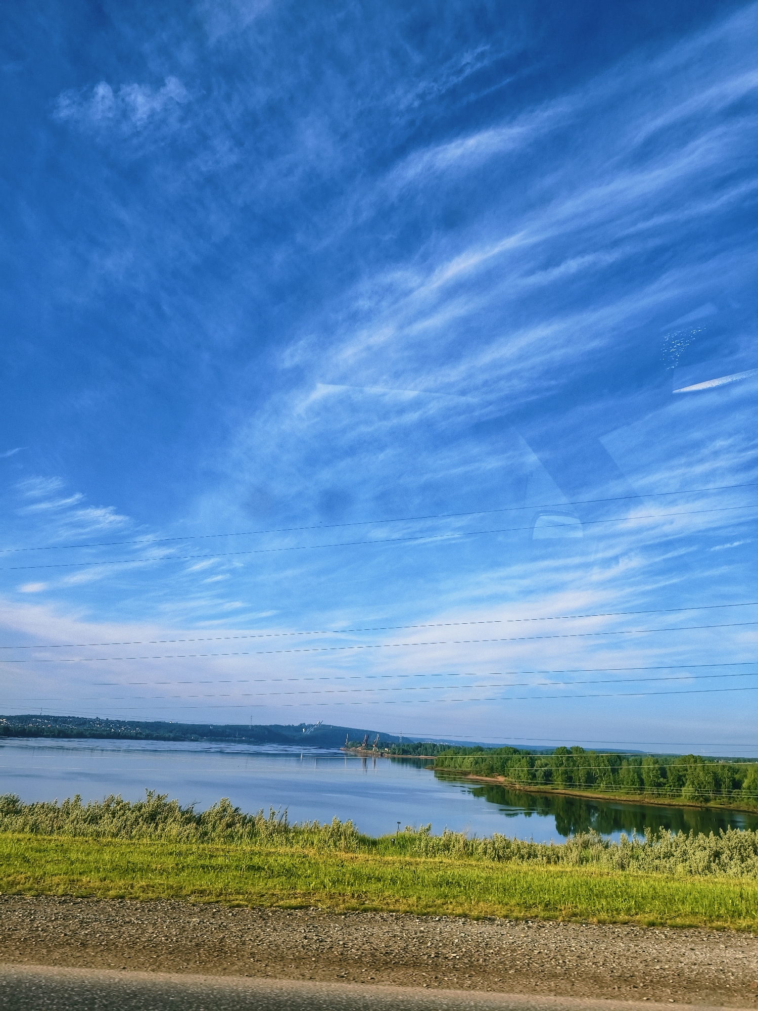 This vertical photograph presents a vast outdoor landscape featuring a wide river under a bright blue sky, taken from what appears to be a moving vehicle or a vantage point near a road. The immediate foreground shows a grassy embankment and a gravel road shoulder.
The central subject is a broad river, its calm surface reflecting the sky. Both the near and distant banks are covered with lush green foliage and trees. On the far left bank, a settlement or town can be seen nestled into distant hills or cliffs. Towards the right side of the far bank, several industrial structures, possibly cranes, are visible.
The sky above is a vibrant blue, adorned with streaky, white cirrus clouds that add texture to the upper atmosphere. Several power lines, some appearing as solid lines and others as dashed segments due to reflections or distortions, horizontally cross the mid-ground and upper sections of the image. Reflections and glare, including what resembles a car's rearview mirror or internal window, are present in the upper right portion of the frame, indicating the photo was taken through a glass barrier.
The time of day appears to be daytime, likely late morning or early afternoon, characterized by clear, bright conditions. The weather is fair. The scene, located near Chaikovsky, Russia, offers a tranquil view of the natural environment combined with subtle indications of human development along the river. There are no people or discernible text visible in the image.