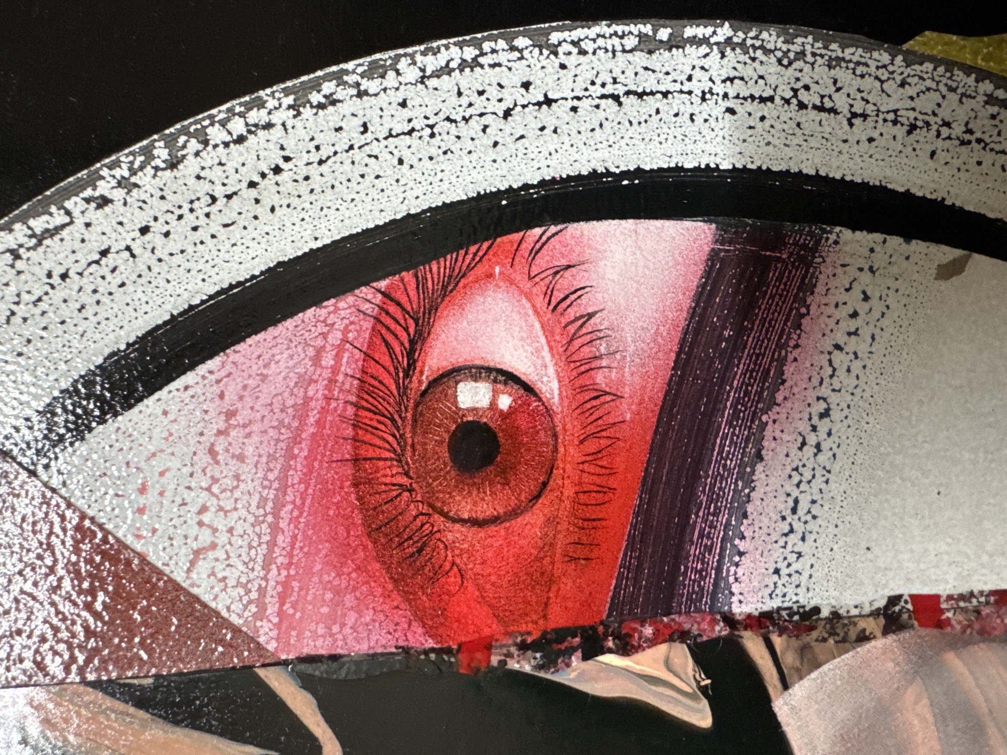 The image displays a close-up of an artwork, featuring a vibrant, stylized human eye with a red iris and pupil. The eye is rendered with thick, dark eyelashes and appears to be part of a larger abstract or graphic design. The artwork incorporates a textured white element with black speckles bordering the eye and a black stripe below it. There are also areas of red and pink gradient. The overall aesthetic suggests a contemporary art piece, possibly a painting or mixed media work. The image does not provide any contextual information about its location in Clonmel, Ireland, or any environmental details beyond the artwork itself.