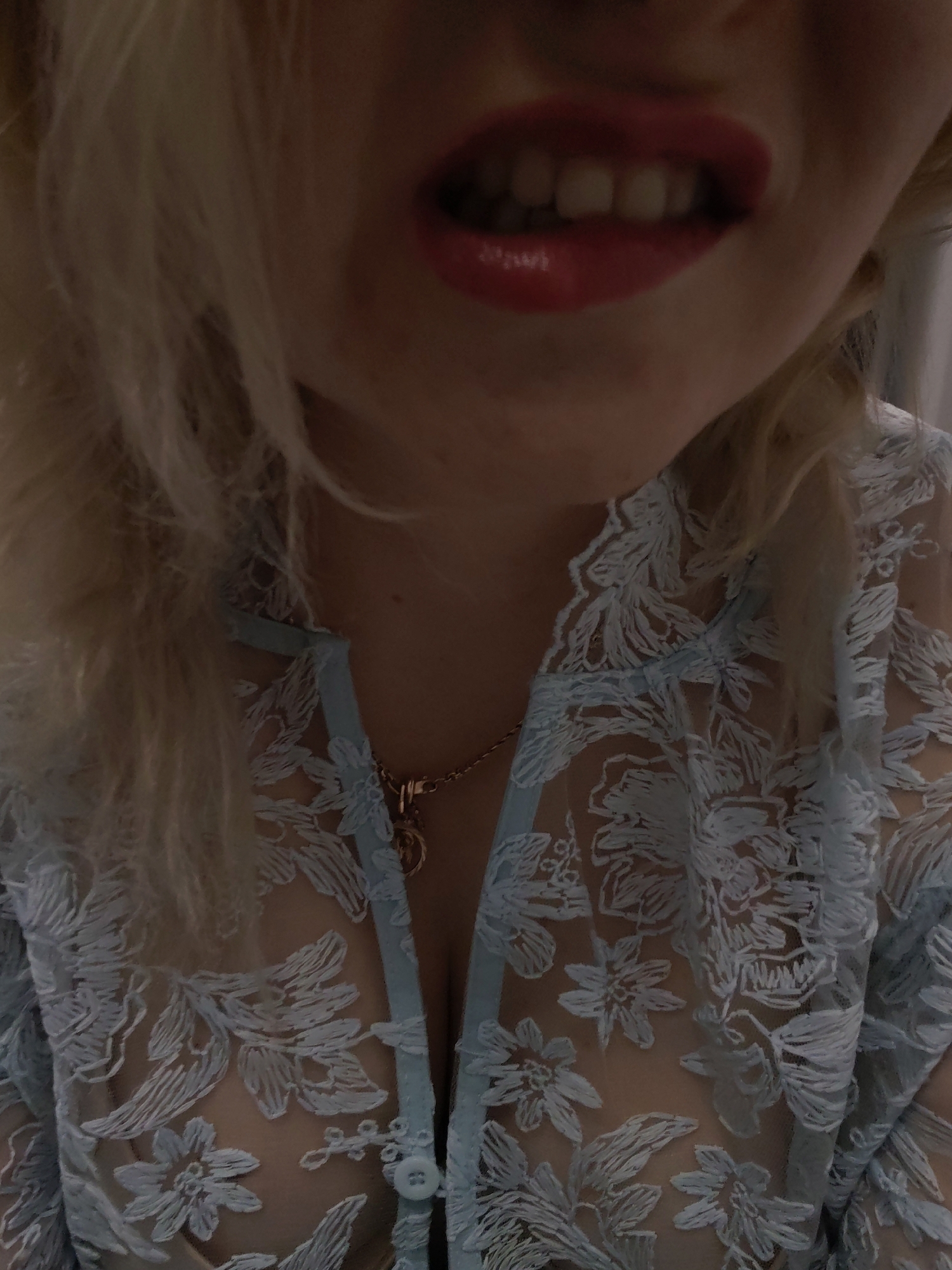 A close-up shot focuses on a woman's face and upper torso. Her light blonde hair is partially visible, framing her face. Her lips are painted a glossy red, and her teeth are slightly visible. She is wearing a light blue, floral lace garment that appears to be a robe or top, partially unbuttoned. A gold necklace with a pendant is visible around her neck. No specific location in Izhevsk, Russia is discernible from the image.
