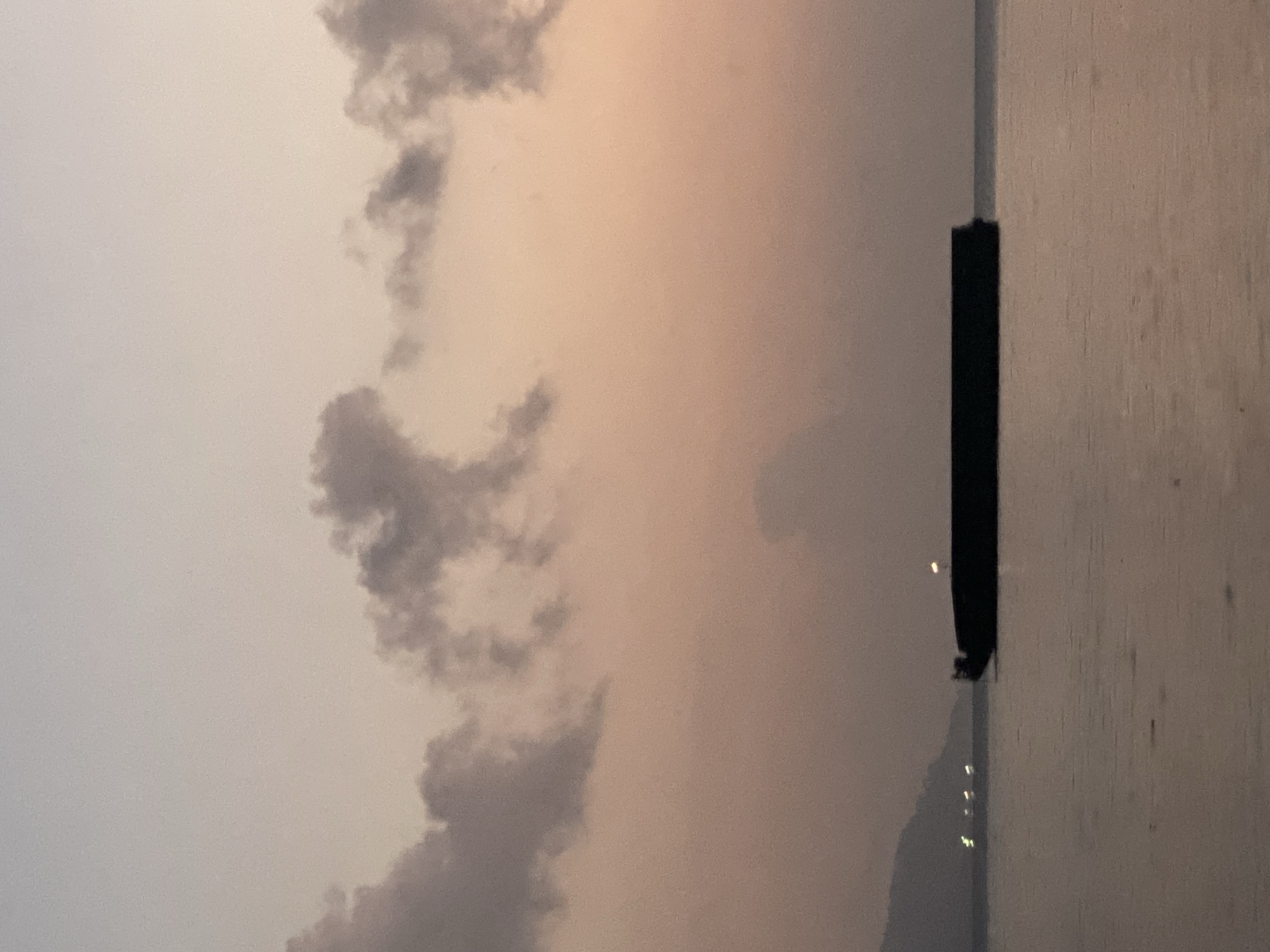 The image captures a serene seascape at dusk near Pulau Bapa, Indonesia. The sky is painted with soft hues of pink and grey, with a few silhouetted clouds drifting across. In the foreground, the calm water reflects the muted colors of the sky. A large, dark barge floats horizontally in the middle ground, its silhouette sharply defined against the horizon. In the distance, the faint outline of a hilly coastline is visible, dotted with a few small lights, possibly from other vessels or structures. The overall atmosphere is one of tranquility and stillness.