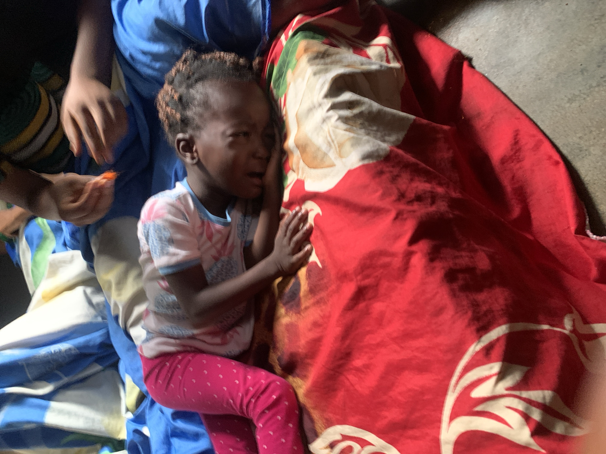 A young child, likely female, is asleep on a bed covered with a red sheet featuring a white and brown abstract design, possibly representing animal features. The child is wearing pink leggings with small white polka dots and a light-colored t-shirt with a patterned design. Their head is resting on their arm, which is on the patterned sheet. To the right of the child, an adult's hands are visible, with the individual appearing to be holding something orange, possibly food. The background shows a blue and white striped sheet or blanket, indicative of a domestic setting in Sabon Tasha, Nigeria. The lighting suggests an indoor environment, possibly during daylight hours.