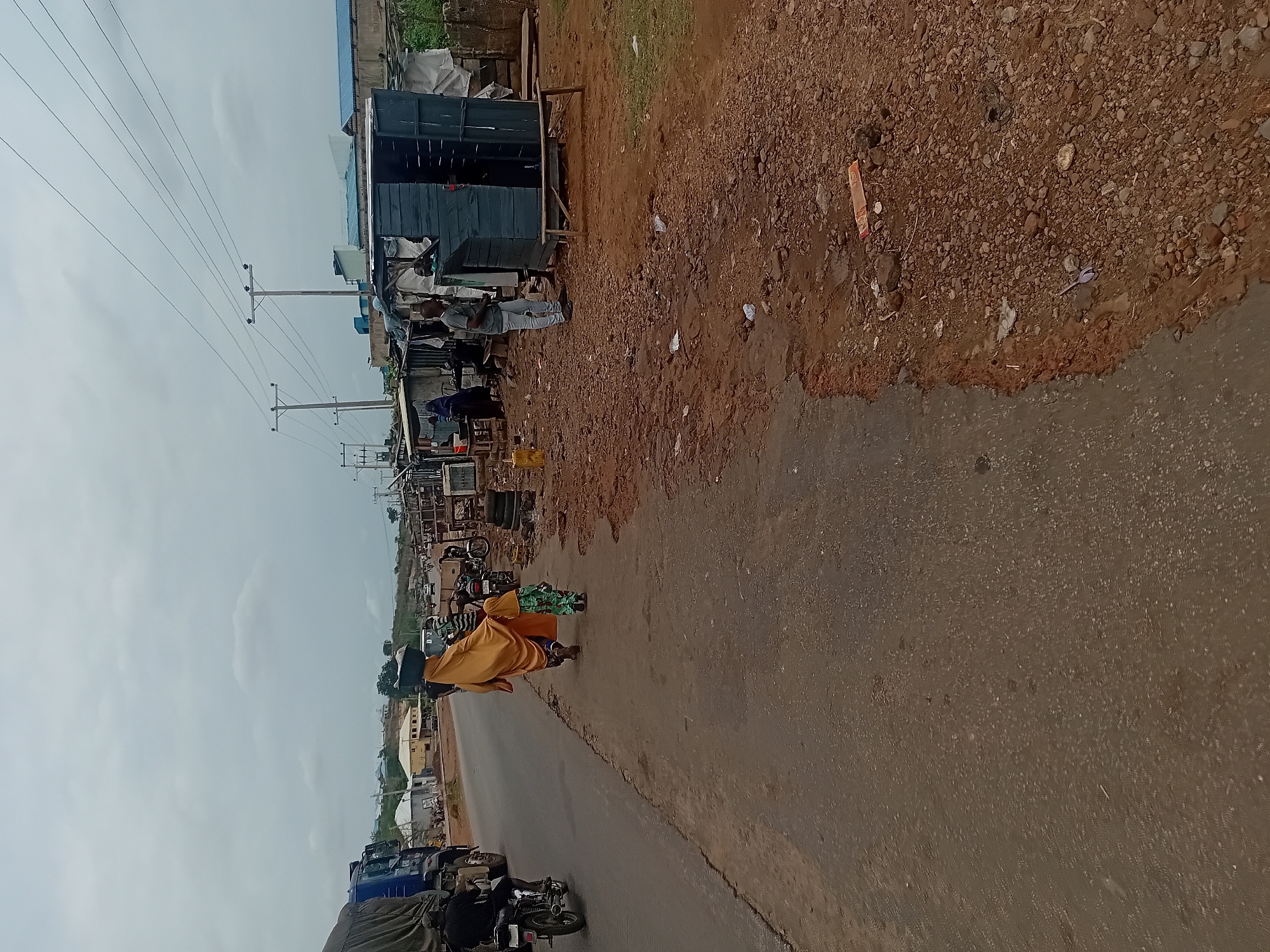 A street scene in an area identified as Unknown, Unknown, shows a paved road running diagonally across the frame. On the left side of the road are several motorbikes and vehicles parked. Further along, there is a line of small stalls and shops, with people standing around or working. One person wearing a yellow garment is walking along the roadside, possibly carrying an object. Overhead are power lines supported by poles. The area next to the road is covered in dirt and small stones. The sky is overcast.