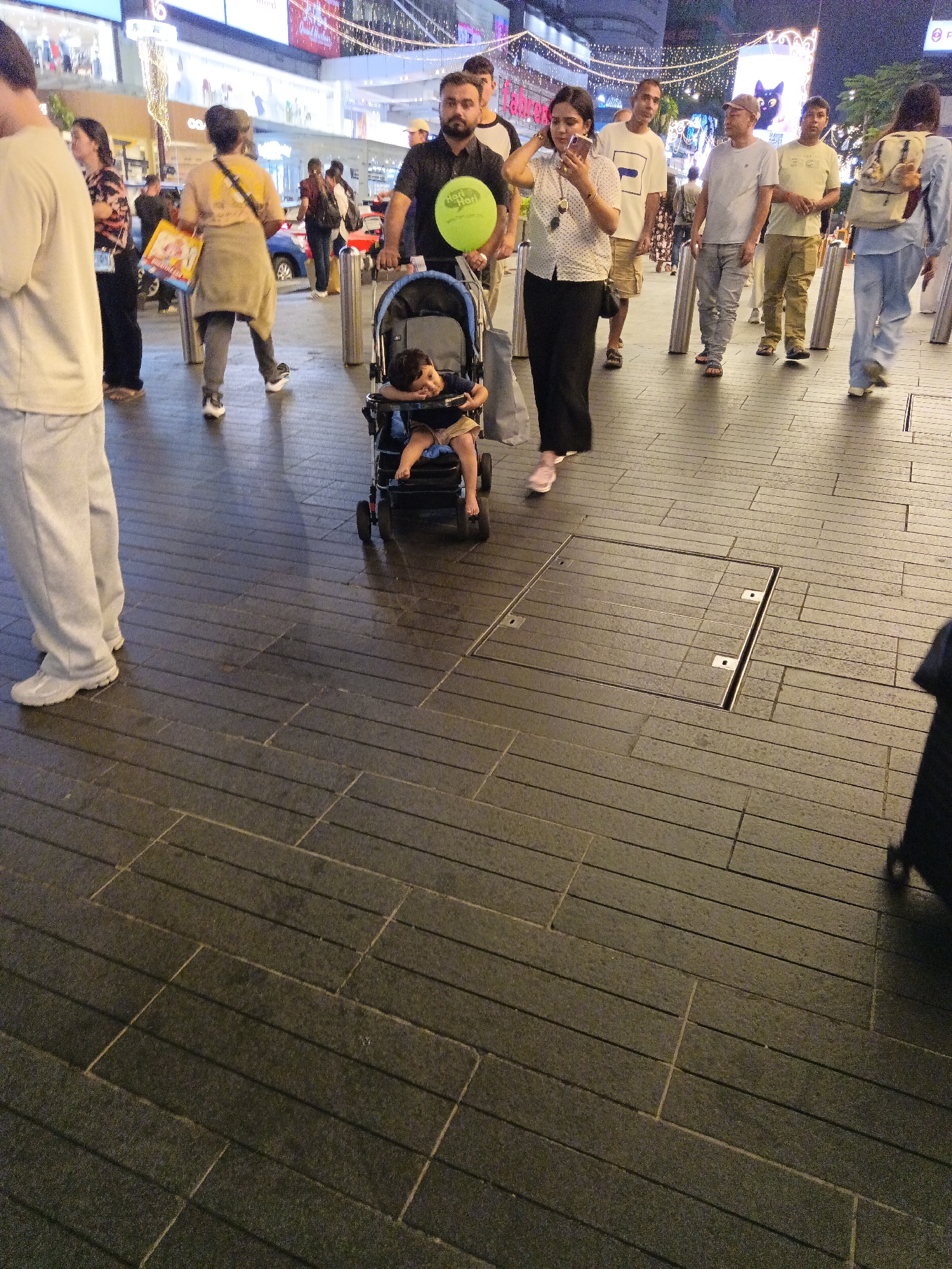 A street scene in Kuala Lumpur, Malaysia, captures a diverse group of people, including families and individuals, navigating a pedestrian walkway at night. The walkway is paved with dark gray, rectangular tiles, with some sections featuring in-laid metal covers. In the foreground, a man pushes a stroller with a child inside. The child is seated and appears to be resting, with bare legs visible. Behind the stroller, a woman in a black dress and a white polka-dot blouse is holding her phone. To the left of the stroller, a man in a light-colored shirt and gray pants is visible. Further back, a group of people walks together, with one woman carrying a yellow bag and another in a light blue outfit with a backpack. The background is illuminated by city lights and signage from shops and businesses, suggesting a bustling urban environment. The overall atmosphere appears casual and social, typical of a public space in a city center.