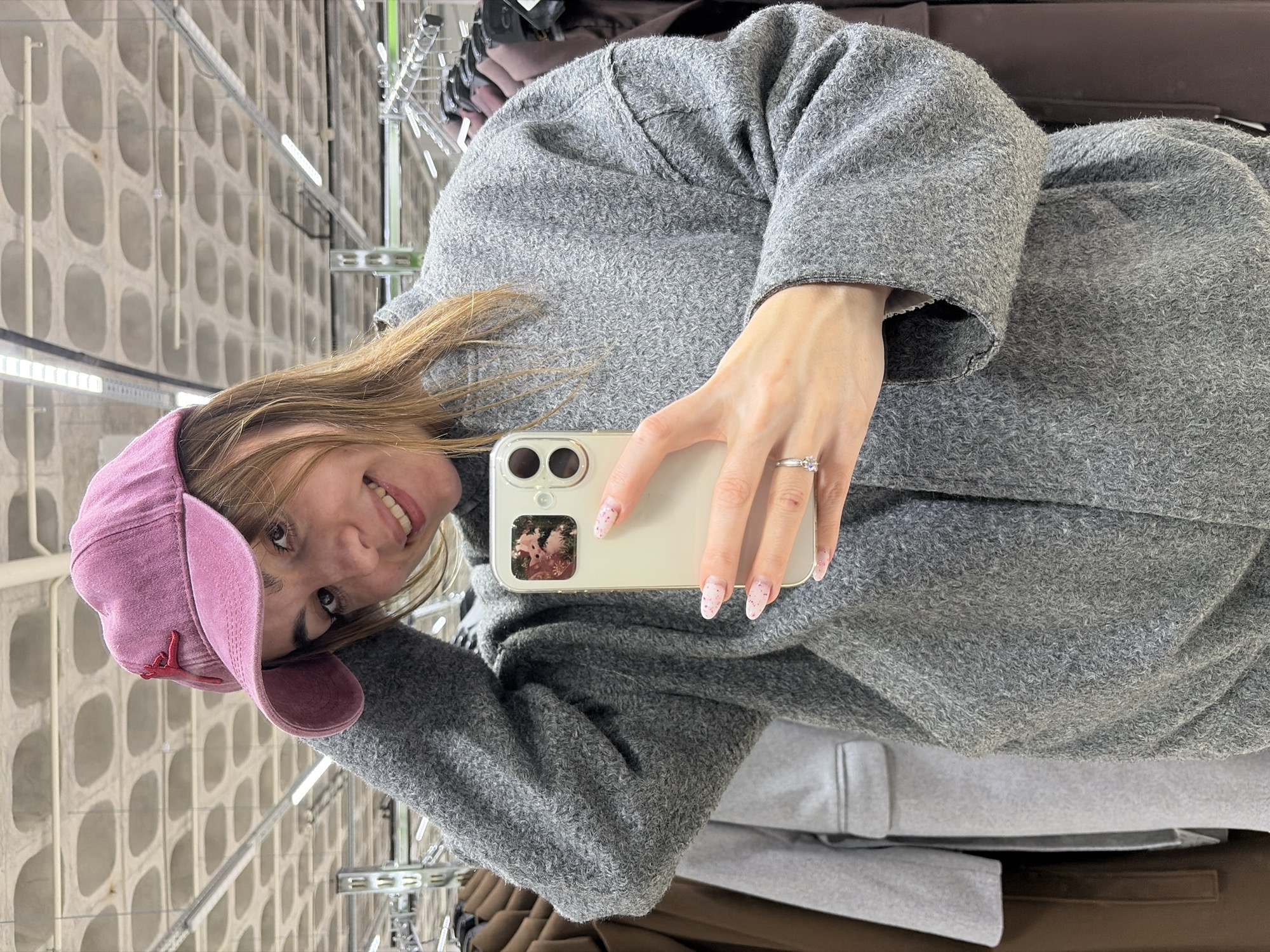 A woman takes a selfie indoors. She is wearing a pink baseball cap and a gray jacket. She is holding a white smartphone in her right hand. Her nails are painted pink with white dots. She is wearing a ring on her right ring finger. Behind her is a concrete wall with oval cutouts. The location could be Izhevsk, Russia.