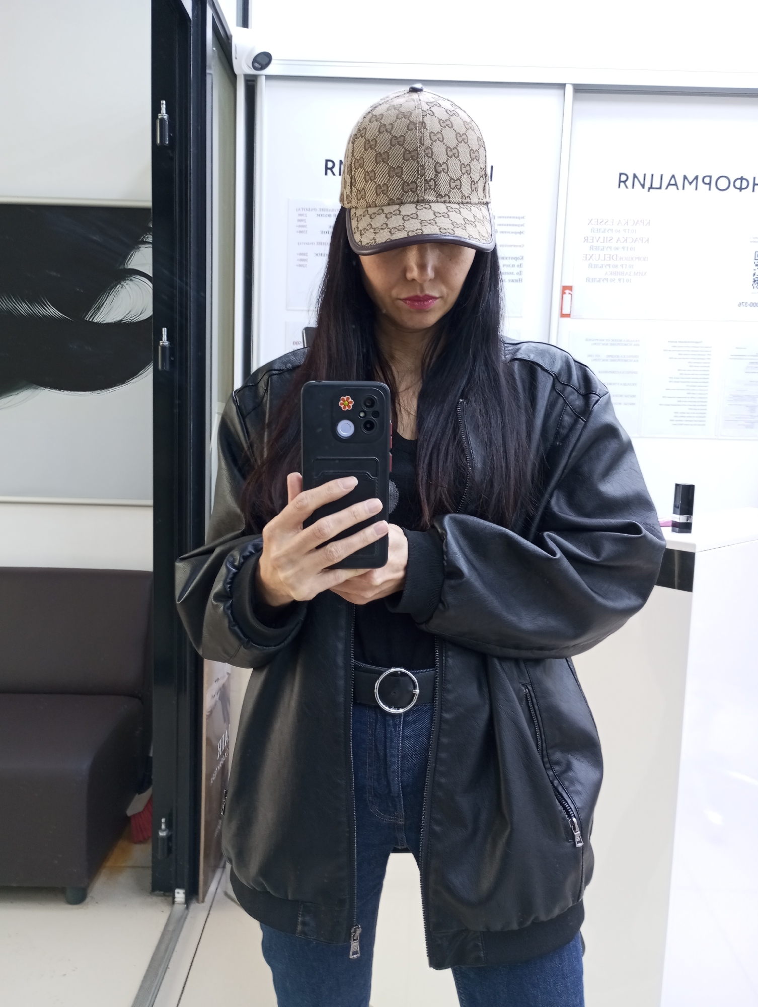 A woman in a Gucci hat, black jacket, and jeans is taking a selfie with her phone in a location identified by background text as being in Izhevsk, Russia. The background contains a wall with text in Russian and what appears to be a security camera mounted above. Other furniture is visible in the background.