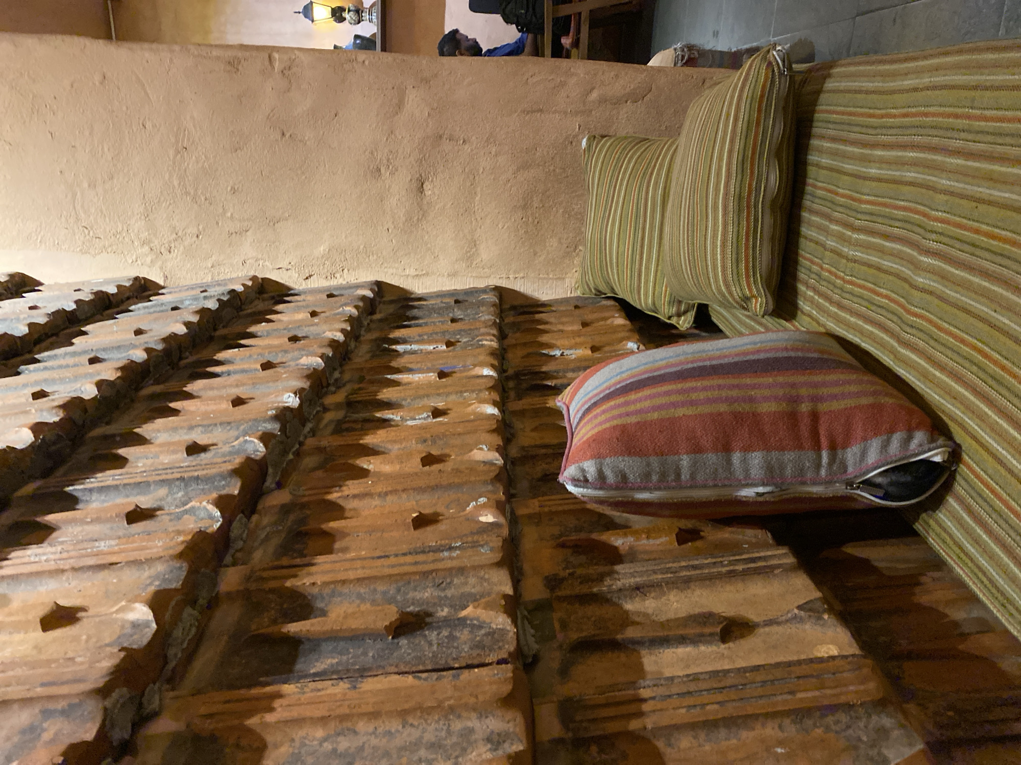 The image depicts an indoor scene with a distinct rustic aesthetic. In the foreground, a bench is covered with a striped cushion and two additional striped pillows. To the left, a wall is constructed from numerous overlapping clay tiles, creating a textured and layered surface. The wall has a warm, earthy tone with visible signs of age and wear. To the right of the tiled wall, a section of a peach-colored, textured wall is visible. Further back, a person is seated at a wooden table, engaged with a laptop. The background includes a shelf with decorative lamps and other objects, suggesting a cafe or restaurant setting. The lighting is dim, contributing to the cozy ambiance. The image does not provide any specific information about the city of Đà Nẵng, Vietnam, or any activities happening in that location.