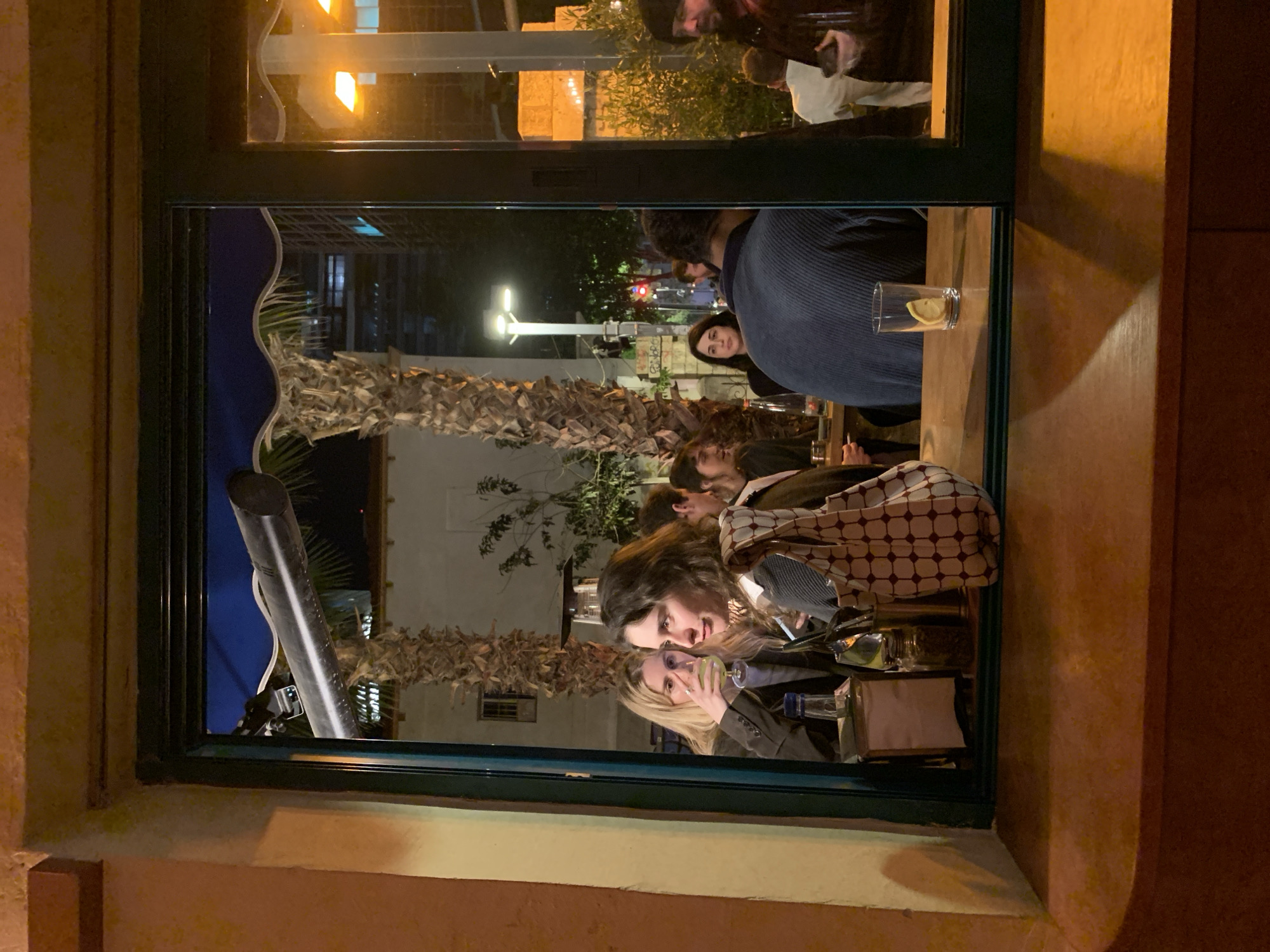 The image depicts a street view of a restaurant in Tel Aviv, Israel, as seen through a window from outside. A group of people are seated at tables inside the restaurant, appearing to be engaged in conversation and dining. Their faces, torsos, and table settings are partially visible. A beverage with a lemon slice rests on a table edge near the window. Outside, part of a palm tree and other urban fixtures are visible. The surrounding architecture suggests a contemporary urban environment. The image is taken at night, illuminated by artificial light from the restaurant and streetlights.