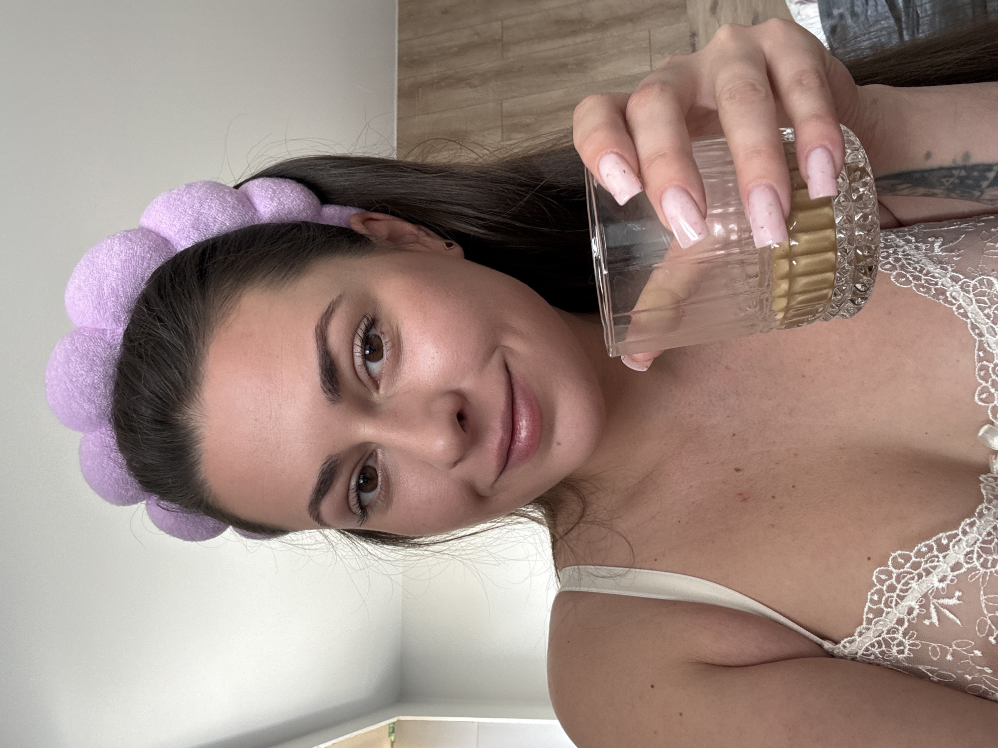 A woman with dark hair pulled back by a purple, puffy headband is holding a glass of amber liquid in her right hand. She is wearing a white lace bralette and is looking towards the camera with a slight smile. The background appears to be an indoor setting with a white wall and wooden flooring. Her fingernails are painted light pink.
