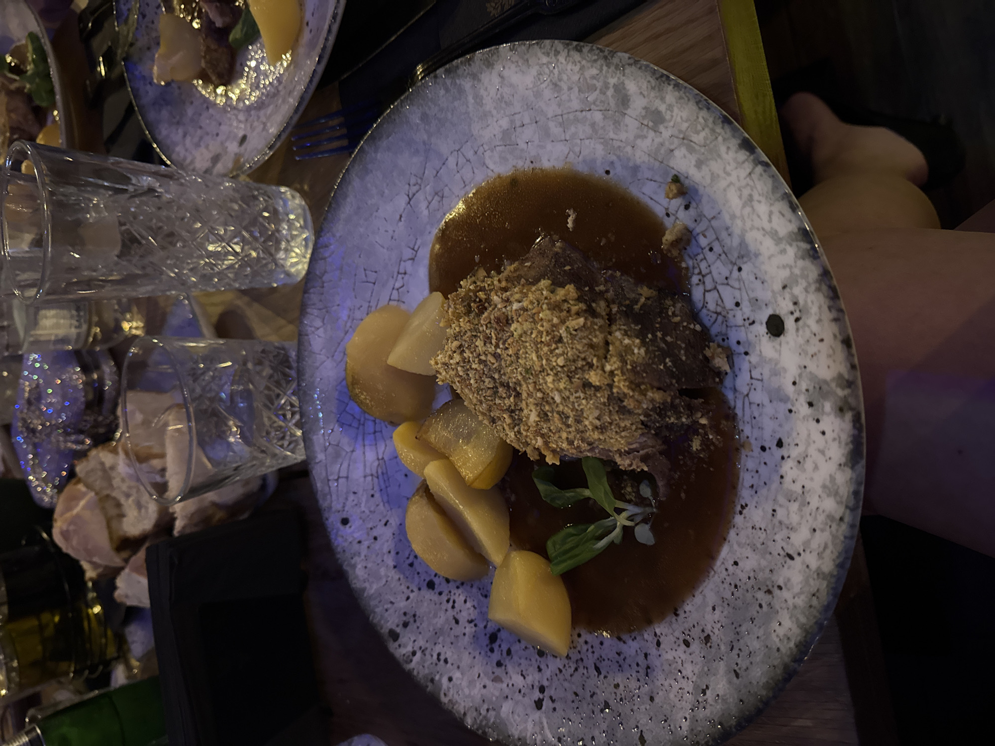 The image shows a top-down view of a plate of food. The plate appears to be made of ceramic and has a mottled gray and white pattern. The food on the plate consists of a piece of meat covered in small, granular crumbs, arranged in a pool of dark brown sauce. Beside the meat are several sliced pears. The plate is sitting on a table. Adjacent to the plate are two glasses and a dish with additional food items. The scene is likely indoors. The image does not provide enough information to determine if it was taken in Blagoevgrad, Bulgaria.