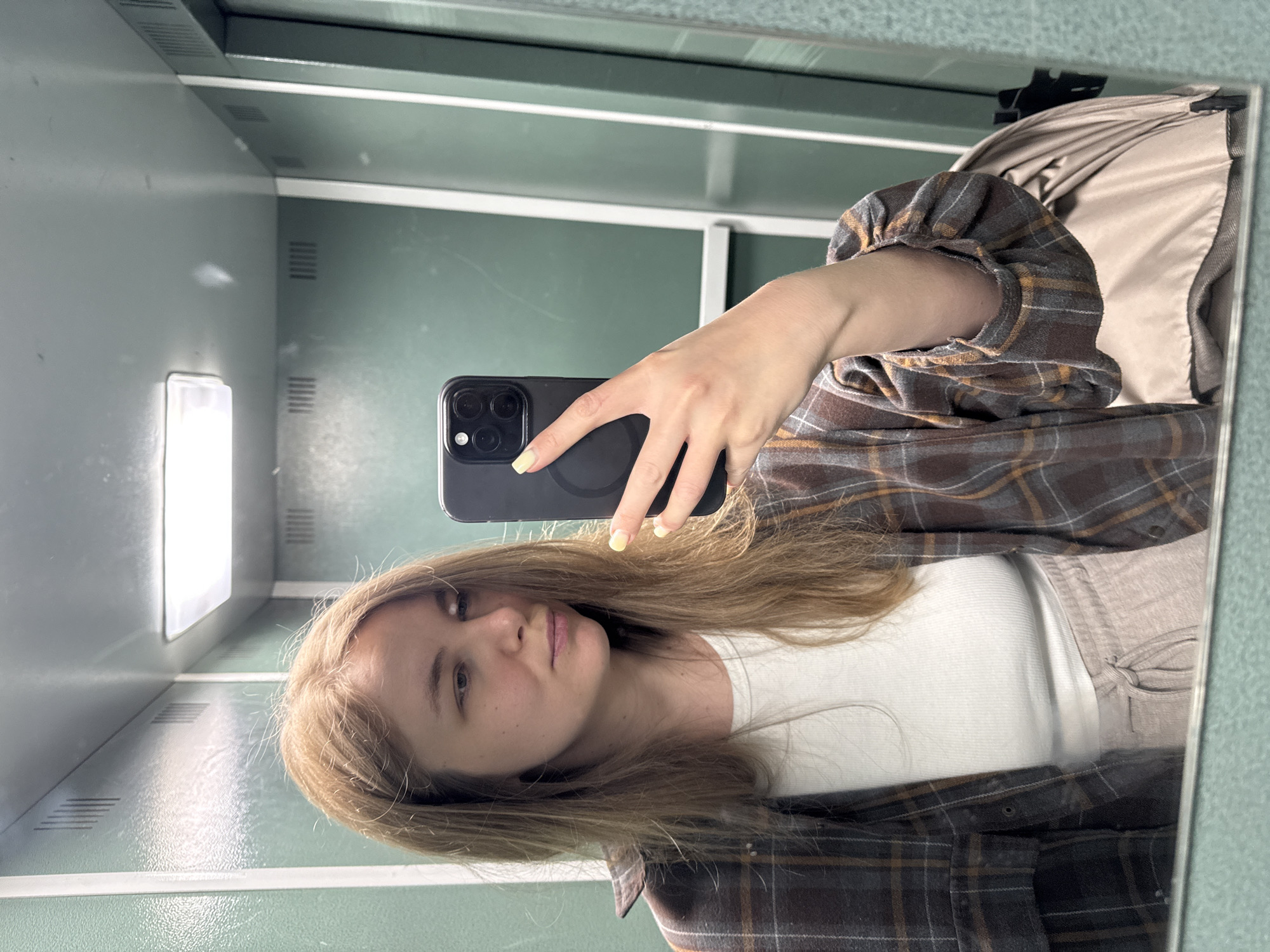 A woman with blonde hair is taking a selfie in a mirrored space. She is holding a black smartphone with multiple camera lenses. She is wearing a white top and a brown plaid flannel shirt. The environment appears to be an institutional setting, possibly a public restroom or changing area, with gray walls, white trim, and artificial lighting. The photo was taken in Vladimir, Russia.